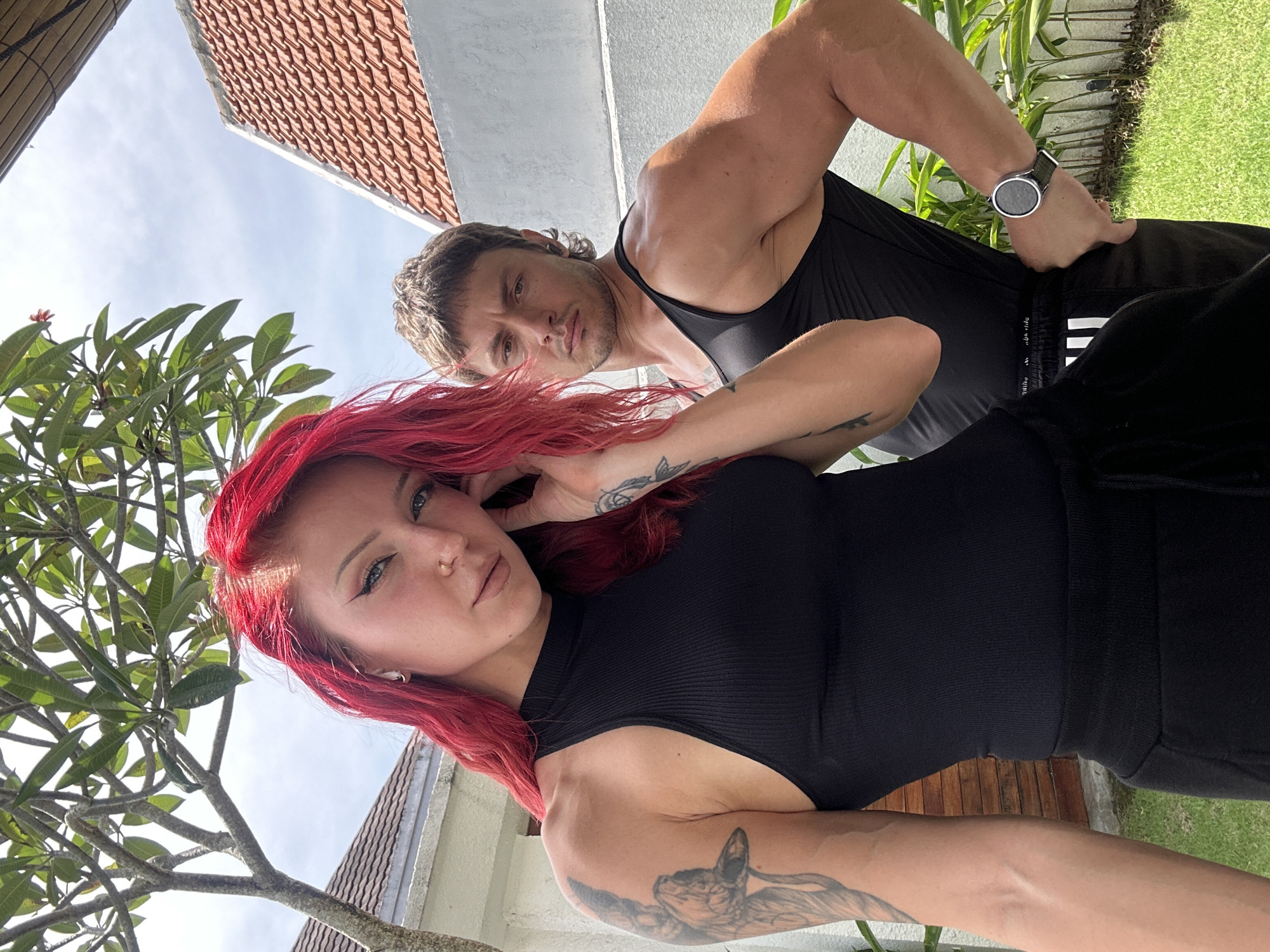 Two people, a man and a woman, are in a selfie. The woman has bright red hair and a black top. Tattoos are visible on her arms. The man is muscular and wears a black tank top and shorts. He has a watch on his left wrist. They appear to be outside; in the background, there is a tiled roof, some greenery, and a section of grass. Based on the information, the image was likely taken in North Kuta, Indonesia.