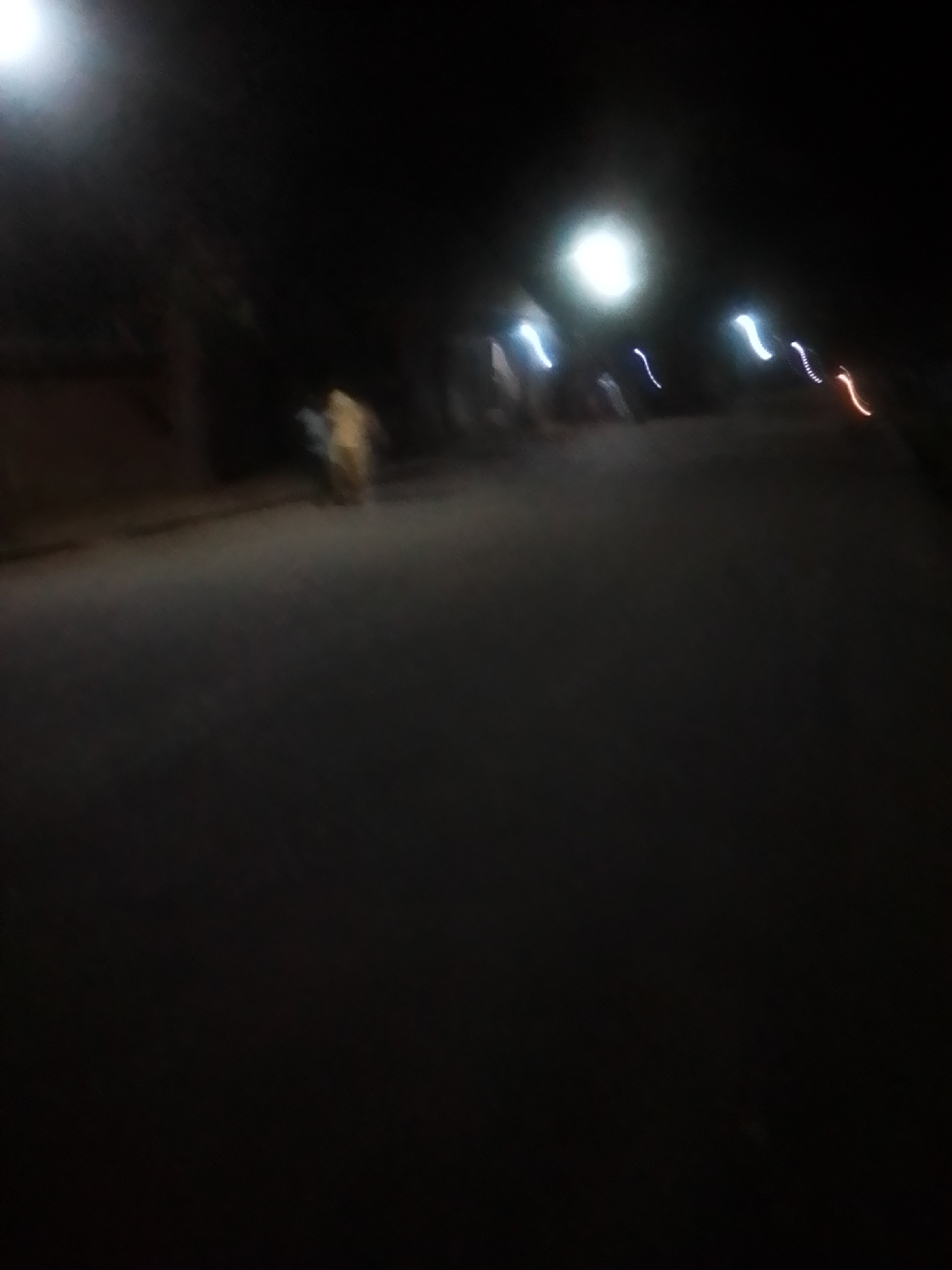 The image depicts a blurry night scene on an asphalt road, presumably in Liji Hill, Nigeria. A bright light source, possibly a streetlight, illuminates the upper left portion of the frame. Streaks of colored lights, likely from vehicles, are visible in the distance, suggesting movement. In the middle ground, two indistinct human figures can be observed. One figure appears to be wearing a light-colored garment, while the other is partially obscured. The overall impression is of a dimly lit street at night, with minimal detail due to motion blur and low light conditions.