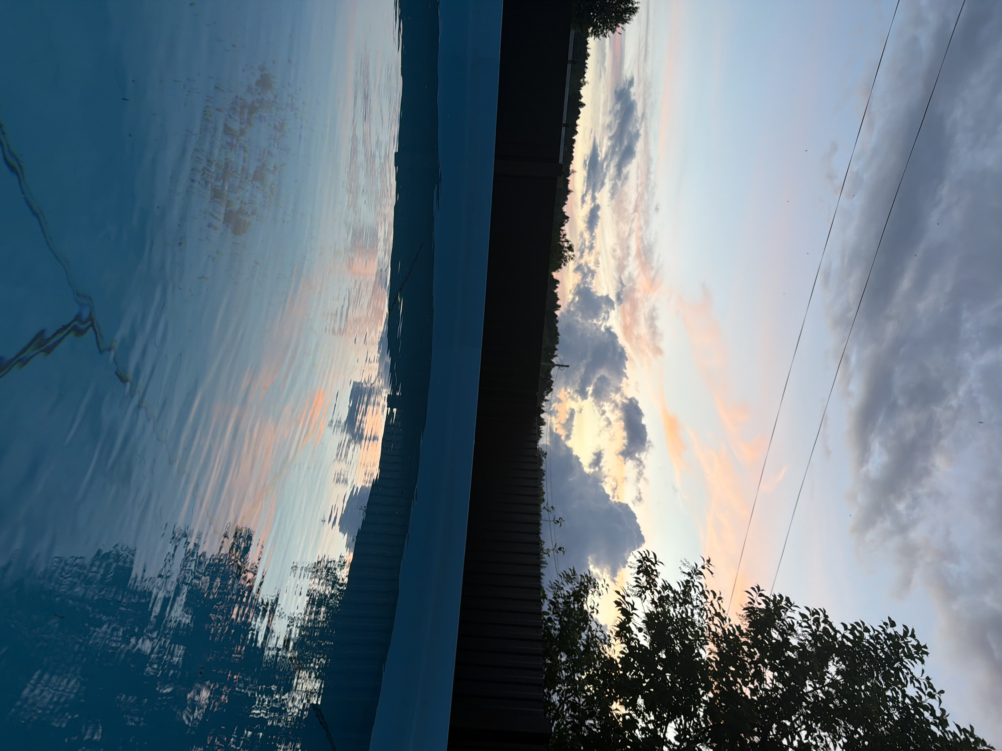 The image depicts a scene at dusk near a swimming pool in Chaikovsky, Russia. The pool water reflects the sky's colors: orange, pink, and blue. A dark fence separates the pool area from a line of trees in the background. A deciduous tree is silhouetted in the foreground. Power lines extend across the sky. The sky shows a mix of clear areas and cloud formations.