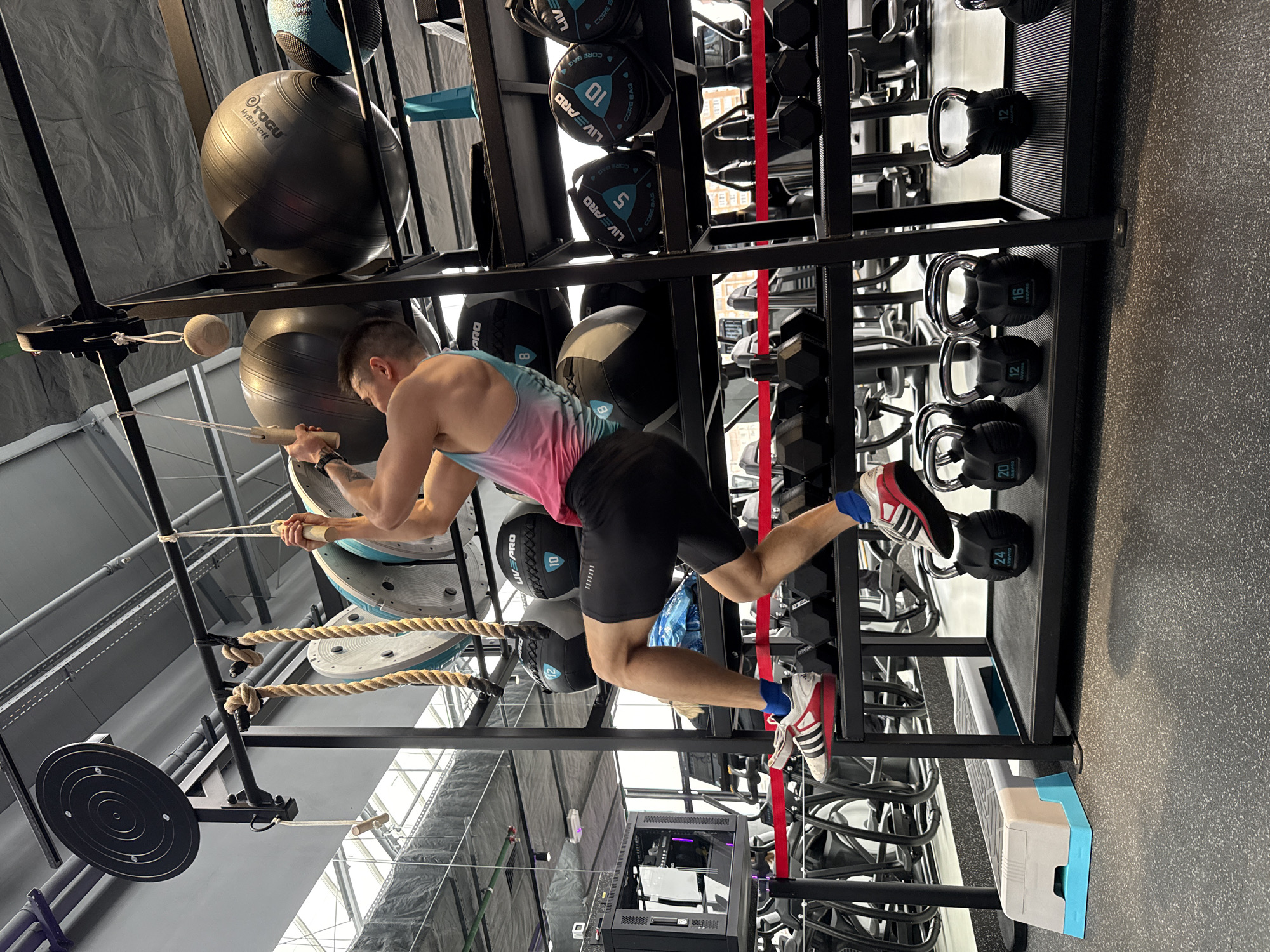 Here is a detailed description of the image:
**Scene Overview:**
The image shows a man exercising in a gym. The gym appears to be well-equipped, with various types of exercise equipment visible.
**People:**
* A man is performing an exercise that involves pulling himself up using ropes or cables. He is wearing athletic clothing (a tank top and shorts) and shoes.
**Objects/Equipment:**
* **Exercise Equipment:** The gym has a variety of equipment including weight racks, medicine balls, kettlebells, dumbbells, and specialized training setups (like the one the man is using).
* **Weight Rack:** A prominent black weight rack holds dumbbells, kettlebells, and weighted balls (some labeled "LIVEPRO CORE BAG").
* **Weighted Balls:** Exercise balls of different sizes are placed on top of each other.
* **Ropes:** There are ropes and cables being used as part of the exercise setup.
* **Gym Flooring:** The floor appears to be a grey gym flooring material.
* **Gym Step:** There's a small grey and blue step in the bottom right corner.
**Action/Interaction:**
* The man is actively engaging in an exercise that appears to require upper body strength. He is pulling himself up using the rope system.
**Environment:**
* **Location:** A well-equipped gym.
* **Lighting:** The gym appears to have adequate lighting, with natural light filtering in from an exterior window or skylight, and artificial lighting from internal fixtures.
* **Layout:** The gym seems organized with equipment neatly arranged.
**Forensic Details:**
* The orientation of the image is rotated.
* The text "LIVEPRO" can be seen on some of the exercise balls.
* The man is wearing shoes with a distinctive red and white striped design.
**Location:**
The location is the city of Kazan, Russia.
