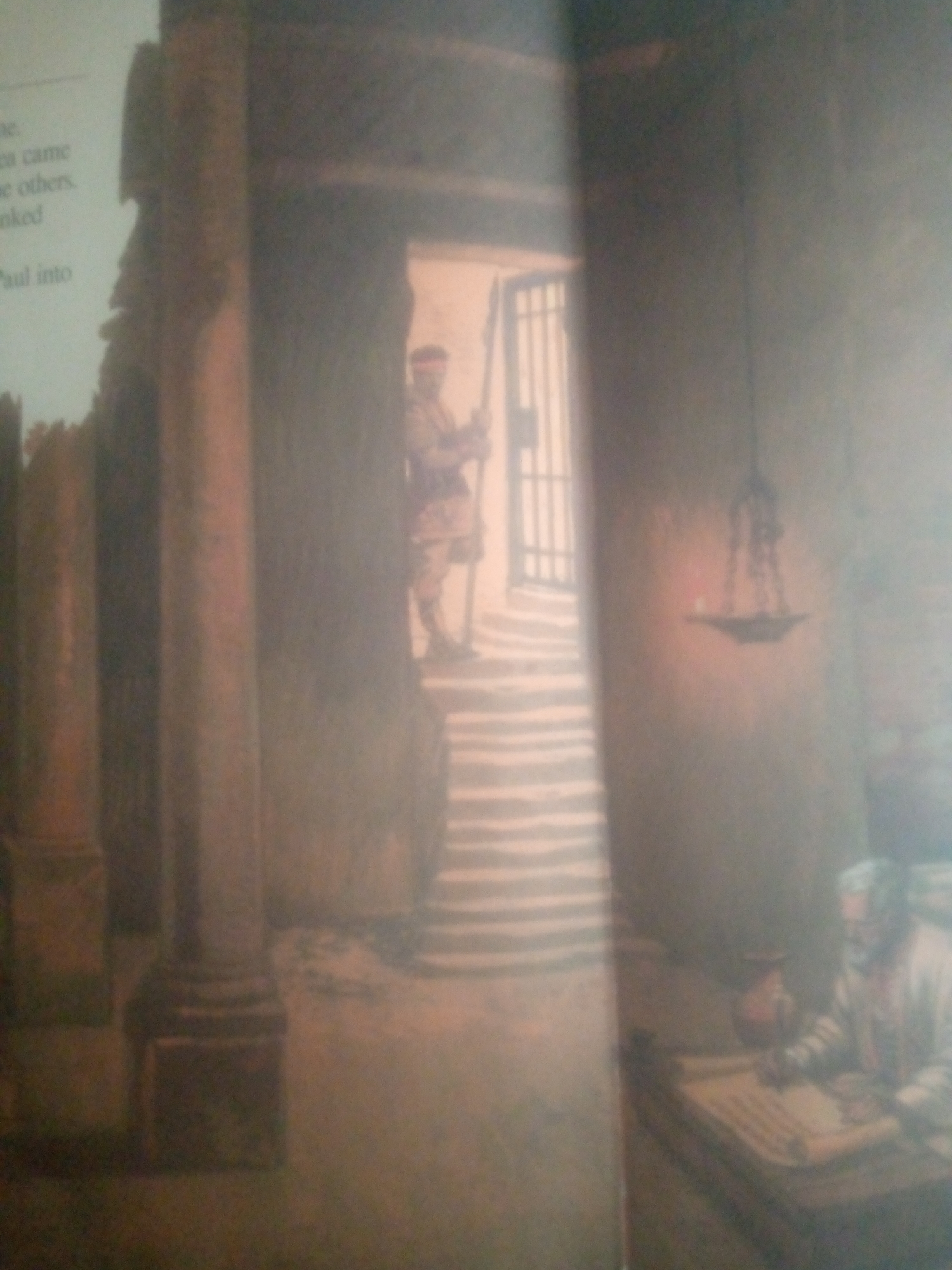 The image depicts an interior scene with a person dressed in historical attire standing in a doorway with a barred gate. The setting appears to be ancient, with stone pillars visible on the left and a staircase leading up to the doorway. The person in the doorway holds a staff or spear. In the foreground on the right, another person, likely a scholar or scribe, is seated at a table with a scroll or manuscript. A lamp hangs from the ceiling. The overall lighting suggests an indoor, possibly dimly lit environment. The text on the left side of the image is partially visible and appears to be from a book or document. The scene does not depict any recognizable landmarks or specific elements that would allow for identification of a location within Jauro Yinu, Nigeria.