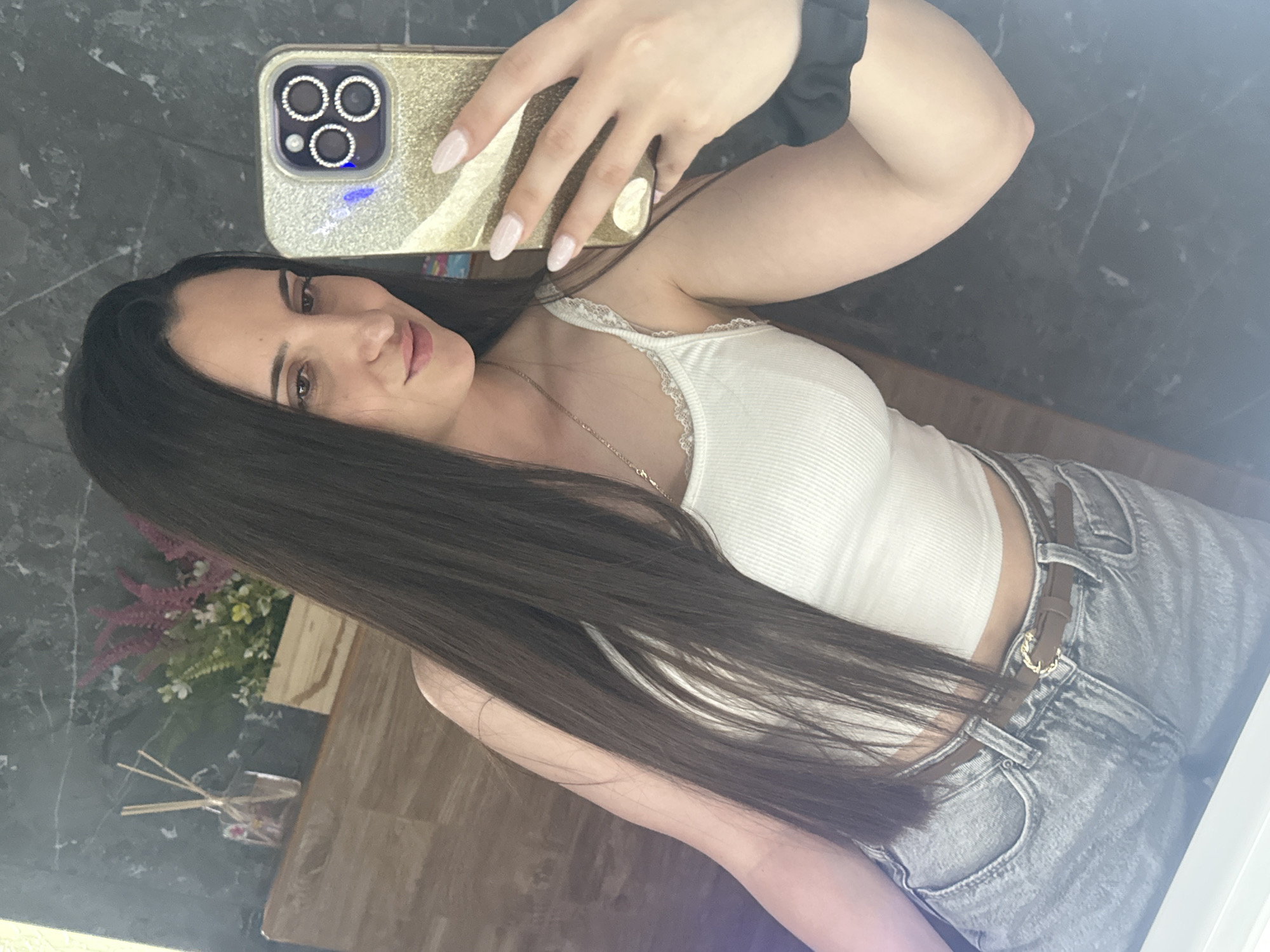 A woman is taking a selfie in what appears to be an indoor setting. The woman has long, dark brown hair and is wearing a white tank top and light wash jeans with a brown belt. She holds a smartphone with a gold case that features decorative rings around the camera lenses. Her nails are long and painted a light color. In the background, there's a gray wall with a marble-like pattern and a wooden surface with a flower arrangement and a reed diffuser. Based on the information provided, it's not possible to ascertain if the photo was taken in Beliakovets, Bulgaria, or any specific location within the city.