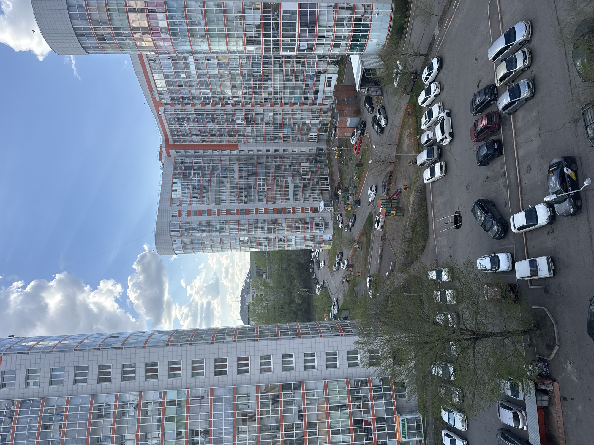 The image shows an aerial view of a courtyard between tall residential buildings in what appears to be Krasnoyarsk, Russia. The buildings are modern, with many windows and some orange detailing. The courtyard is paved and contains parking areas filled with cars, as well as trees, walkways, and a small playground. Some people are visible on the walkways. The sky is blue with white clouds.