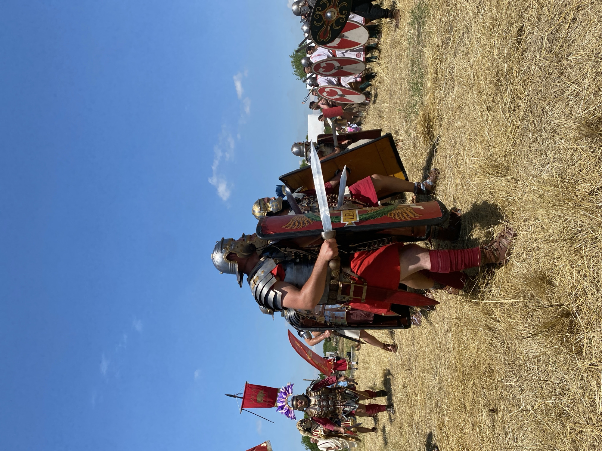 The image depicts an outdoor historical reenactment of Roman soldiers in Gigen, Bulgaria, under a clear blue sky with minimal white clouds. The ground is covered in dry, yellowish-brown straw and sparse grass, suggesting an arid, open field environment.
In the immediate foreground, slightly right of center, stands a man dressed as a Roman legionary. He wears a silver-colored galea helmet with prominent cheek guards and a dark beard visible beneath. His torso is covered by a lorica segmentata (segmented plate armor), and he wears a red tunic or skirt (focale) and red leg wraps, with brown caligae (military sandals) on his feet. He holds a gladius (short sword) in his right hand, angled upwards, and a rectangular scutum (shield) in his left hand, adorned with red and gold details, including part of the word "LEG". He is looking towards his right.
Behind him and to his left, a second Roman soldier is partially visible, similarly armored and also holding a shield and sword. To the left in the mid-ground, a group of figures advances. A prominent figure wears a bronze helmet with a large, elaborate purple plume, a decorated cuirass, and a red tunic. Behind him, another individual wears an animal pelt over their shoulders. Further left, a red vexillum (banner) bearing white text, partially visible as "LEGA", flies from a pole.
To the right in the mid-ground, a line of approximately ten or more additional figures, also in Roman-style attire, stands or marches. Many wear white tunics and carry round shields, predominantly white with red concentric circles or patterns. One shield on the far right is black with intricate golden scrollwork. Some figures in this line also wear helmets and are armed with spears or pila.
In the far background, faint outlines of trees or vegetation are visible, along with a white tent or structure. The overall scene captures a staged procession or formation of Roman military reenactors.