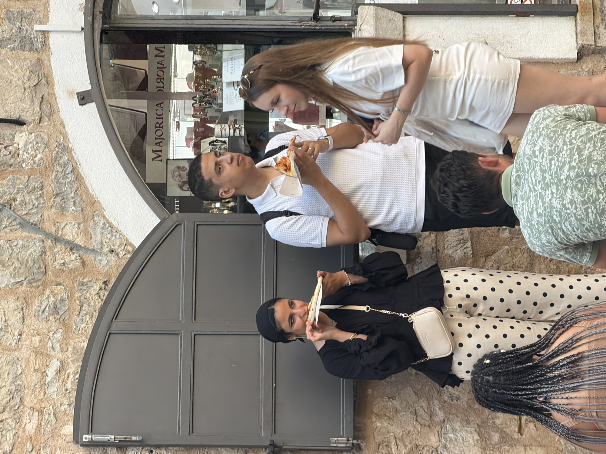 In Budva, Montenegro, a group of people are gathered outside a shop. A woman in a black headscarf and black top with polka dot pants is eating a slice of pizza. Beside her, a man in a white polo shirt is also holding a slice of pizza. To his right, a woman in a white dress stands with her hand on her hair. In the foreground, a person with braided hair is visible from behind, and another person in a green patterned shirt is also in the foreground facing away from the camera. The background shows a stone wall, a dark grey door, and a shop window displaying jewelry. The overall scene suggests a casual outdoor gathering in a European city.