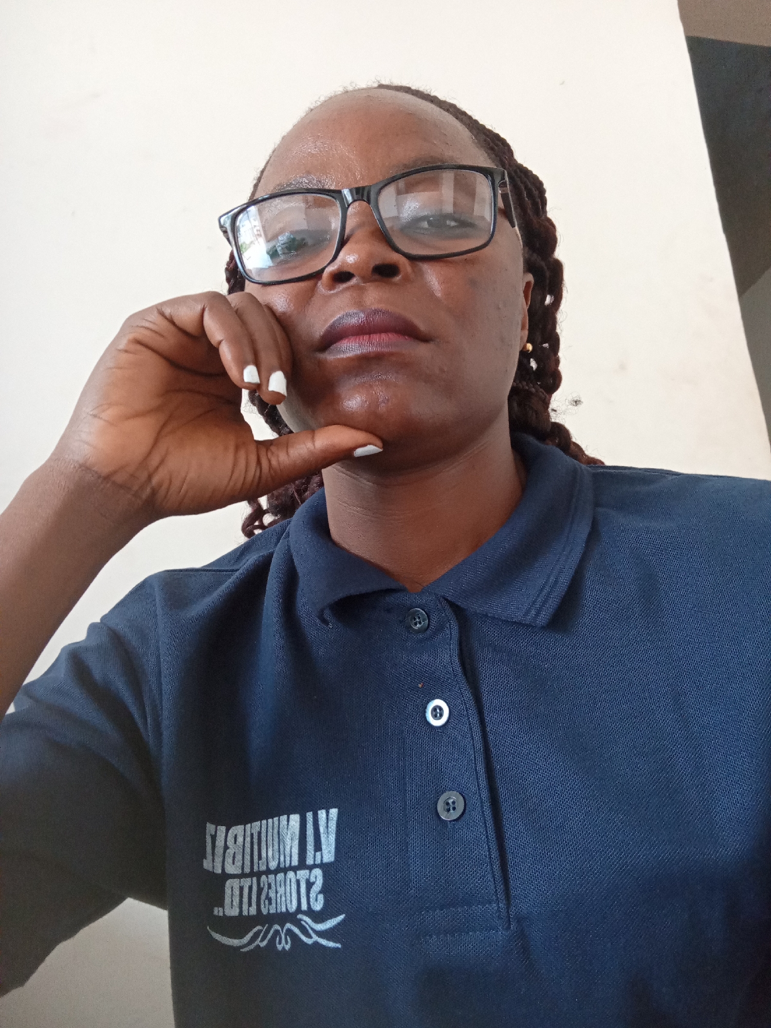 This is a medium shot, head and shoulders, selfie of a Black woman wearing glasses and a dark blue polo shirt. She is looking directly at the camera with a neutral expression, her right hand resting on her chin. Her hair is styled in braids and her fingernails are painted white. The polo shirt has a logo on the left side of the chest that appears to be white text and some decorative swirls. The text is partially obscured by the collar and folds of the shirt, but visible portions suggest it might read "IV STADIUM" or similar. The background is a plain, light-colored wall, with a darker angled surface visible on the right side of the frame, suggesting an indoor setting. The lighting appears to be natural and somewhat harsh, creating slight reflections on her glasses. The scene suggests a casual, everyday moment, likely captured indoors during the daytime in Jalingo, Nigeria, given the provided location context. There are no indications of specific weather or time of day beyond general daylight.