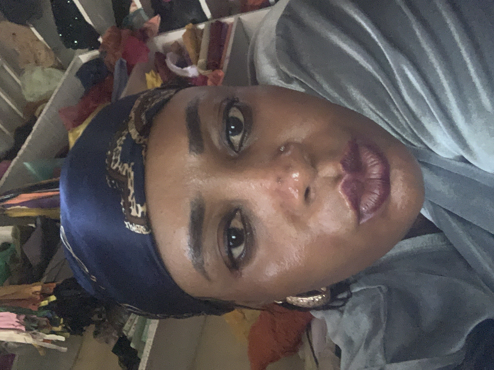 A close-up, slightly angled portrait of a person with dark skin and brown eyes. The person is wearing a patterned blue headscarf and a light gray garment. The person has a nose ring, defined eyebrows and lips. There appears to be some minor acne on their face. The background shows a cluttered storage area with various fabrics and garments. There is no indication in the image that the location is in the city of Sabon Tasha, Nigeria.