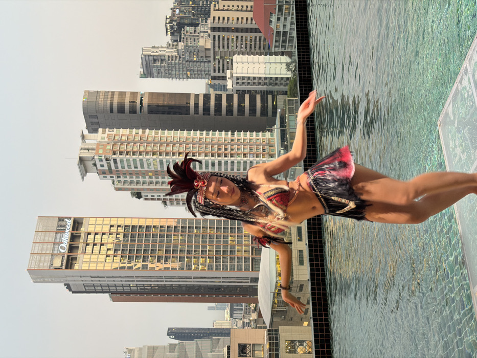 The image displays a female individual in an elaborate costume, standing on a clear platform situated over an infinity-edge rooftop swimming pool. The individual is positioned centrally, with arms extended outwards and slightly upwards, hands open in a dynamic pose. Her head is tilted downwards.
She wears a prominent red feather headdress with an ornate, beaded headband. Her hair is styled in dark braids, potentially with extensions, cascading over her shoulders. The costume includes a red, black, and gold patterned bikini-style top with intricate embellishments, a short, black feathered or fringed skirt with red accents, and various necklaces and arm adornments.
The pool water is clear, appearing turquoise, with visible surface ripples. A dark tiled edge borders the pool.
The background presents a panoramic view of a dense urban environment under a light, overcast sky. Numerous high-rise buildings populate the cityscape. A tall, golden-hued skyscraper on the left side of the frame clearly displays the "Oakwood" logo near its summit, identifying it as the Oakwood Hotel & Residence Bangkok. This structure helps confirm the location as Bangkok, Thailand, within a high-density district such as Sathorn or Silom. Other modern buildings of varying architectural styles complete the metropolitan backdrop.