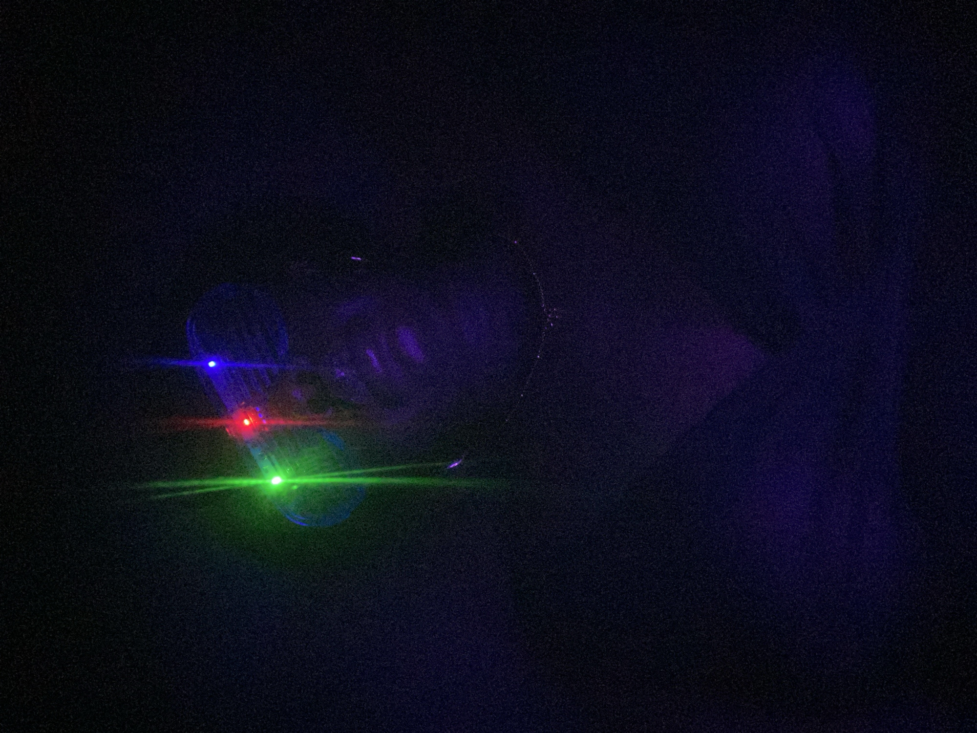 The image depicts a dark environment with a person wearing glasses and multiple laser pointers reflecting off the lenses. There are green, red, and blue laser beams visible. The person's face is partially obscured and appears to be in shadow. The location, Maigizo, Nigeria, is not evident from the image.