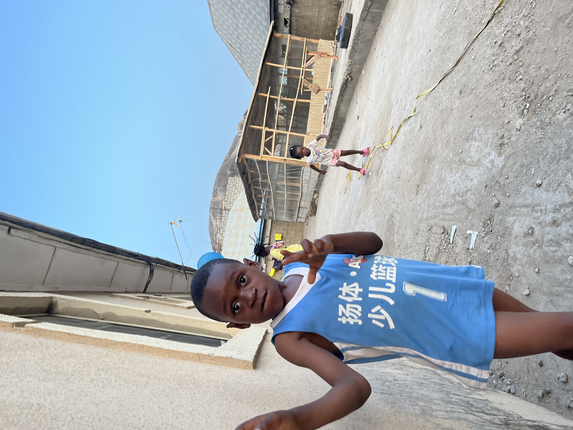A young boy wearing a blue basketball jersey with white trim and Chinese writing, and the number "1", stands in the foreground, extending one hand towards the camera in a "peace" gesture. He is positioned close to a light-colored, one-story building with a window. Behind him, further down a gritty, concrete yard or alleyway, is another child with pigtails wearing a light-colored dress, with pink shoes, who appears to be walking near a yellow cord or rope. Behind the second child is a crudely constructed wooden structure with a roof, and partially covered with sheet metal or some similar material. The scene takes place in Iddah, Nigeria, under a clear, blue sky. The environment appears to be a residential or mixed-use area of the city.