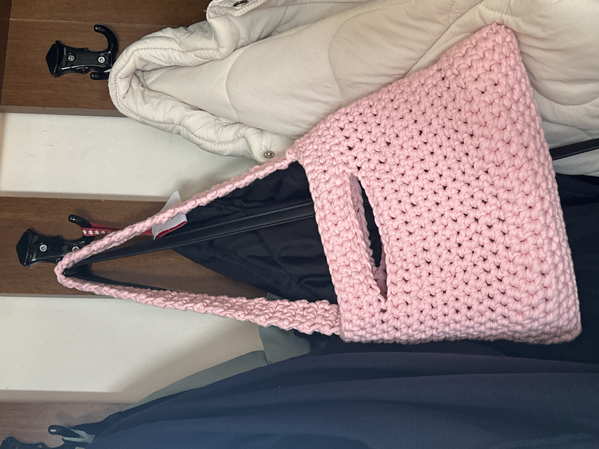 A crocheted pink tote bag with a cutout handle hangs from a black hook on a wood-paneled wall. The bag is positioned in front of a black jacket and to the left of a cream-colored puffer jacket. Other black coat hooks are visible above and to the left of the main subject. The background is a light-colored wall. The scene appears to be an indoor coat rack area.