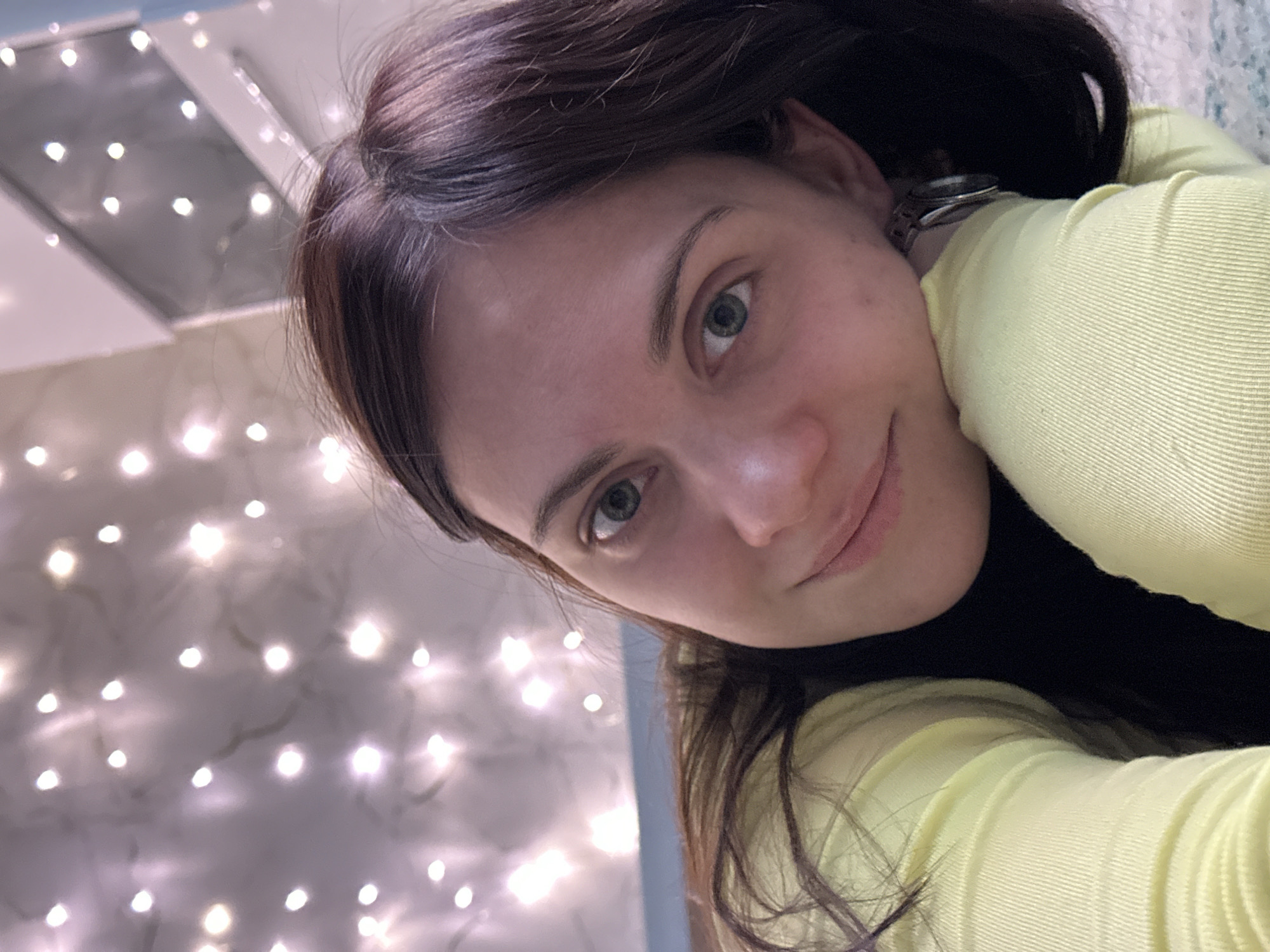 A selfie shows a woman with fair skin, brown hair, and green eyes. She is wearing a light yellow long-sleeved shirt. A glimpse of a silver-colored wristwatch is visible on her wrist near her ear. The background consists of a wall adorned with string lights, and a window frame is visible in the top left corner of the image. The setting is consistent with an indoor environment, possibly a home or apartment. There's no verifiable way to confirm the location as Chaikovsky, Russia from visual data provided.