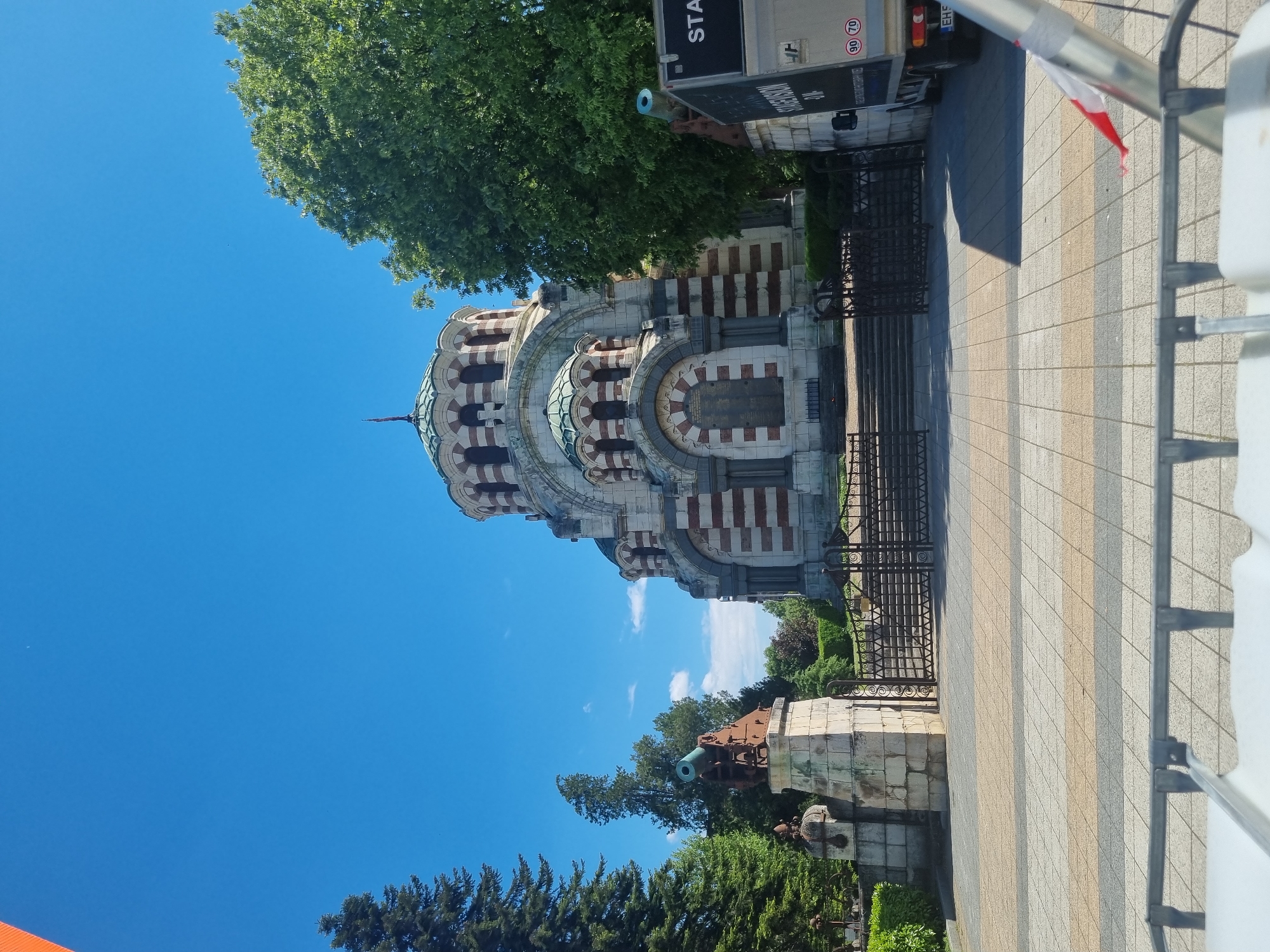 The image depicts the Saint George the Victorious Chapel Mausoleum in Pleven, Bulgaria. The building is a large, ornate structure with white stone and red brick stripes. A dome tops the chapel. Iron fences surround the building. In front of the chapel is a stone monument featuring a cannon. A truck is parked to the right of the chapel. The sky is blue and clear, with trees and greenery framing the scene. A paved walkway with gray and tan striped tiles leads up to the chapel. A white, metal structure is visible to the right of the walkway.