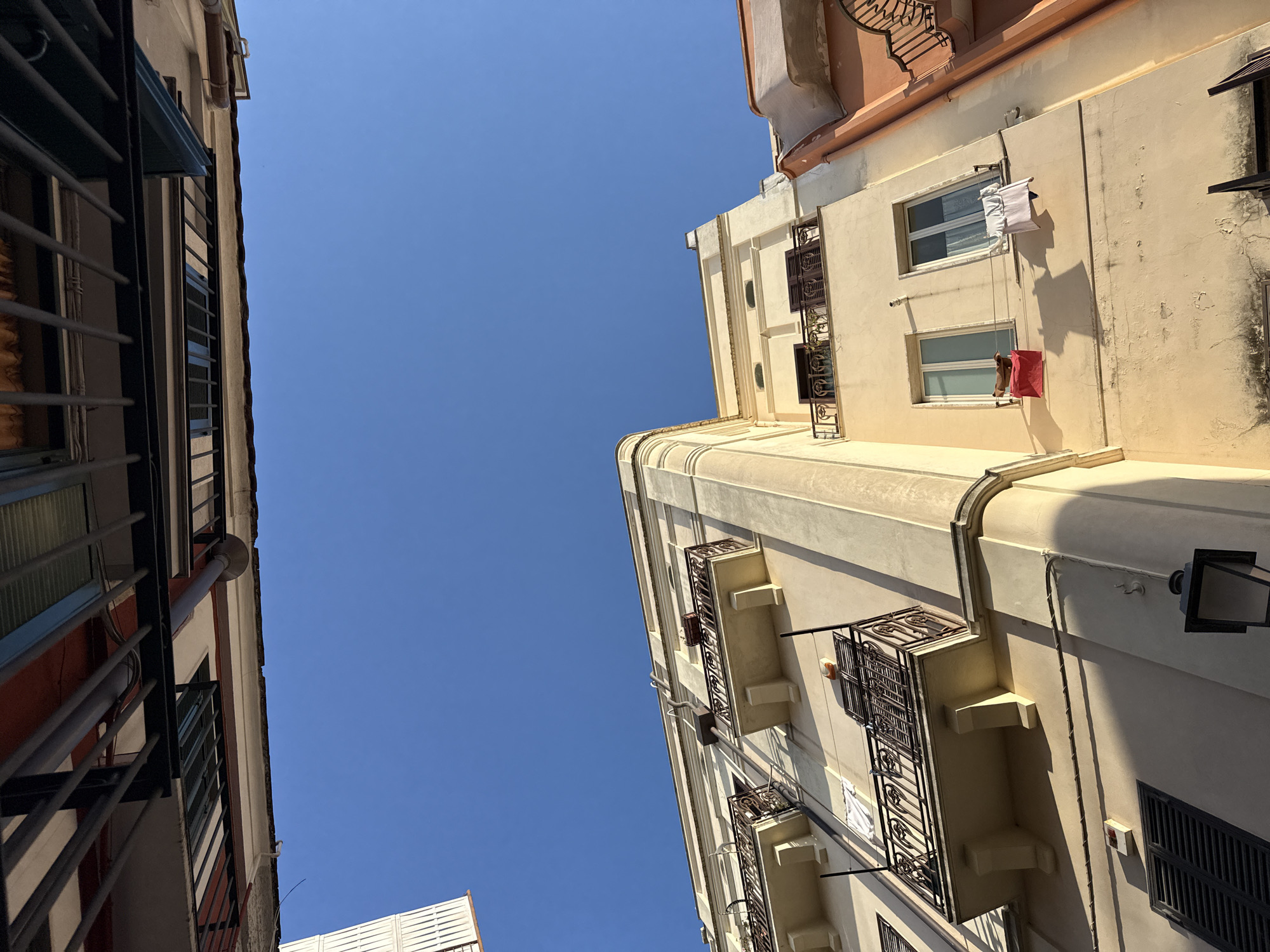 The image presents an upward-looking view between two urban buildings, framing a clear, vibrant blue sky. The perspective is tilted, creating a dynamic composition with the structures leaning inwards from the sides.
On the right, a prominent building with a creamy yellow or beige facade dominates the frame. It features multiple levels with various windows, some equipped with dark louvered shutters, others showing interior blinds. Several balconies, adorned with intricate black wrought iron railings, extend from the building. Notably, a white cloth or garment hangs from a line outside one window, and a red bag or piece of fabric is similarly suspended from another, suggesting laundry drying in the sun. The building exhibits rounded architectural details on its upper sections and visible utility pipes running along its exterior.
A section of another building is visible on the left, characterized by a reddish-brown wall and dark metal balcony railings or window grates. Similar utility conduits are also present on this structure.
The sky, occupying a significant portion of the image, is a uniform, cloudless blue, indicating clear weather. Strong, direct sunlight casts sharp shadows across the facades of both buildings, suggesting it is mid-day or early afternoon. The architecture, including the style of the balconies and facades, is typical of many urban areas in Mediterranean or Southern European regions. No people are visible, and there is no discernible text within the image. The scene captures a slice of urban life under a bright, sunny sky.