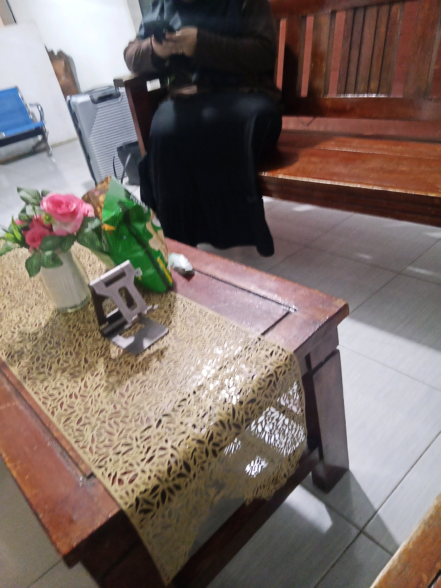 In an indoor setting, a wooden bench is positioned against a wall. In front of the bench, a rectangular wooden table holds a vase with pink roses, a silver phone stand, and a green bag of chips. A person wearing a dark hijab and dark clothing is seated on the bench, holding a mobile phone. To the left of the bench, a silver suitcase stands upright next to a blue chair. The floor is tiled with light-colored square tiles. The scene suggests a waiting area, possibly at an airport or bus terminal, given the presence of luggage. The specific location within Lubuklinggau, Indonesia, is not discernible from the visual evidence alone.