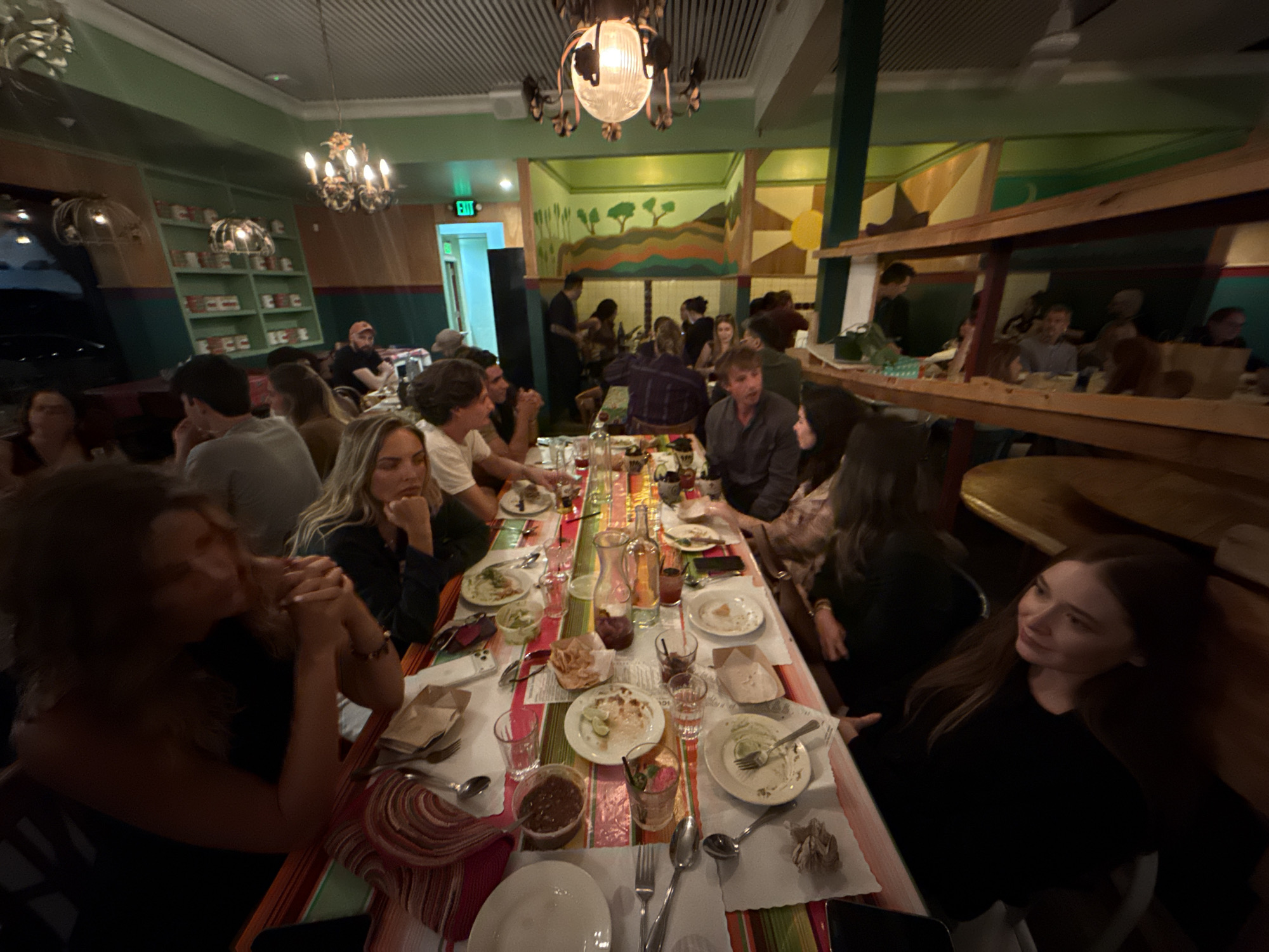 The photograph depicts a group of people dining at a restaurant. The location is likely in Los Angeles, United States of America.
The scene shows a long table covered with a colorful striped tablecloth, laden with plates, silverware, glasses, and various food items. A group of individuals, including men and women of varying ages, are seated around the table.
Some people are engaged in conversation, while others appear to be eating or drinking. The restaurant has a warm and inviting atmosphere, with unique interior decor including shelves of canned goods, chandeliers, and a mural. Additional diners and staff members are visible in the background.