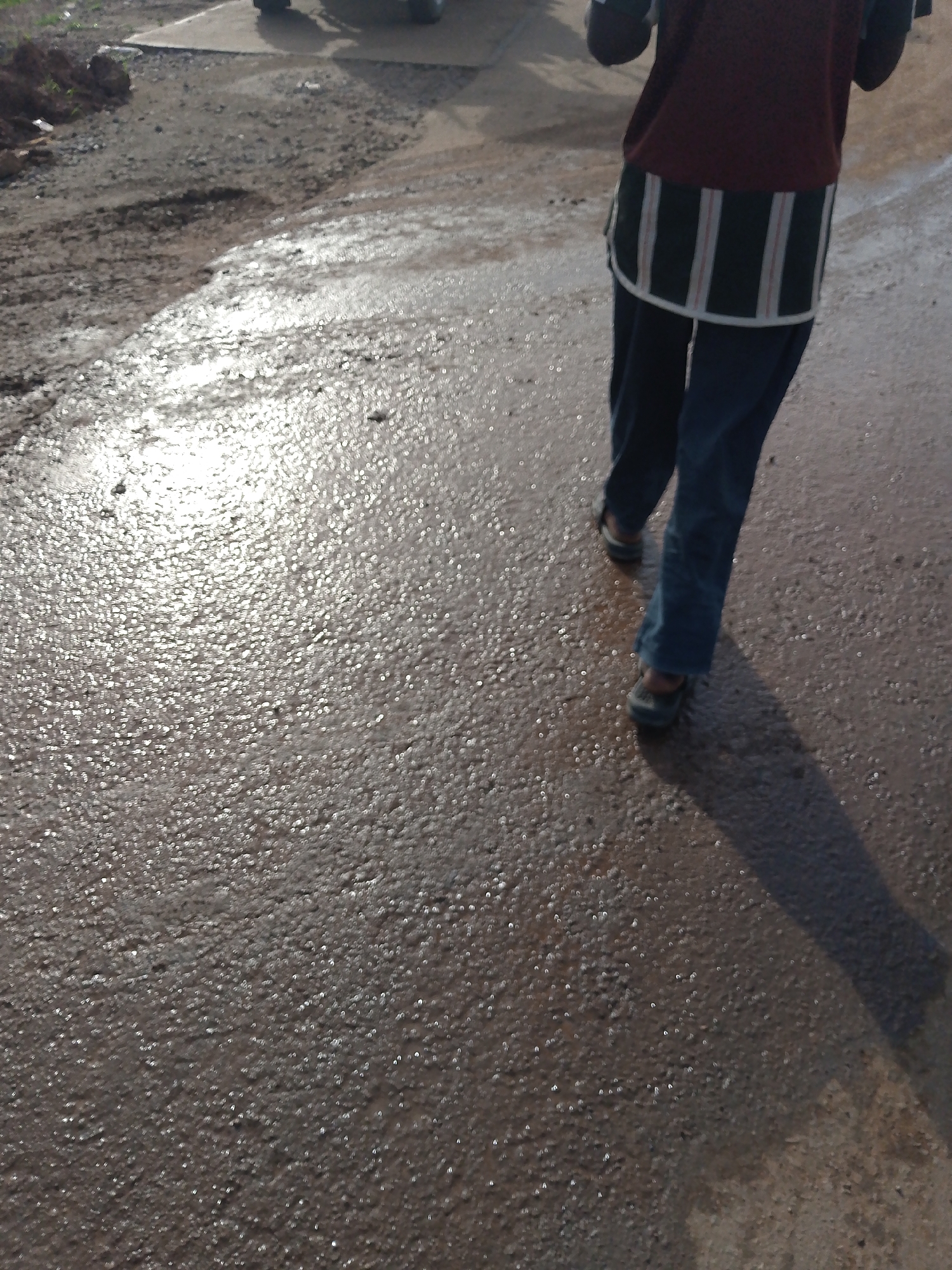 The image shows the lower body of a person walking on a wet, uneven dirt and asphalt surface in Keffi, Nigeria. The person is wearing dark blue pants and a maroon shirt with a striped fabric overlay that has vertical white and dark green stripes. They are also wearing dark shoes. The ground is illuminated by sunlight, creating reflections and highlights on the wet surface. The background shows a pile of dirt and some gravel. A shadow of the person is cast on the ground. The scene appears to be an outdoor area, possibly a road or pathway, that has recently been wet, perhaps from rain or water runoff. The overall impression is one of a person traversing a damp, rural or semi-urban environment.