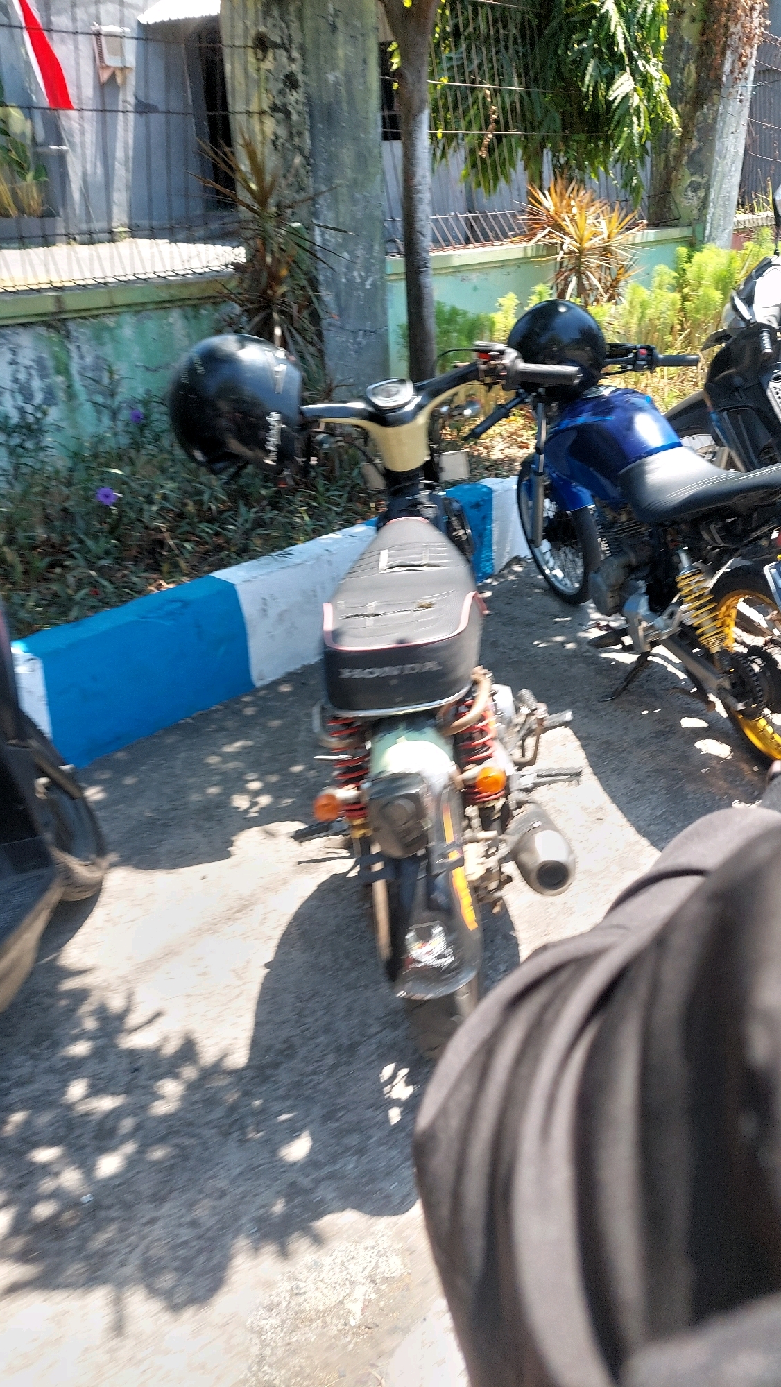 In Siwalan Panji, Indonesia, two motorcycles are parked on a paved surface, possibly a sidewalk, bordered by a blue and white concrete barrier. The motorcycle in the foreground is a light beige and black scooter with a black helmet placed on its handlebars. It features red shock absorbers and an orange turn signal. The motorcycle behind it is a blue and black standard motorcycle with yellow accents on its wheels and a black seat. A black fabric object, potentially clothing, occupies the bottom right corner of the frame, obscuring part of the scene. The background includes a building with a white wall and a red and white flag, alongside a wire fence and various plants and trees, suggesting an outdoor setting under sunlight, evidenced by the distinct shadows cast on the ground.