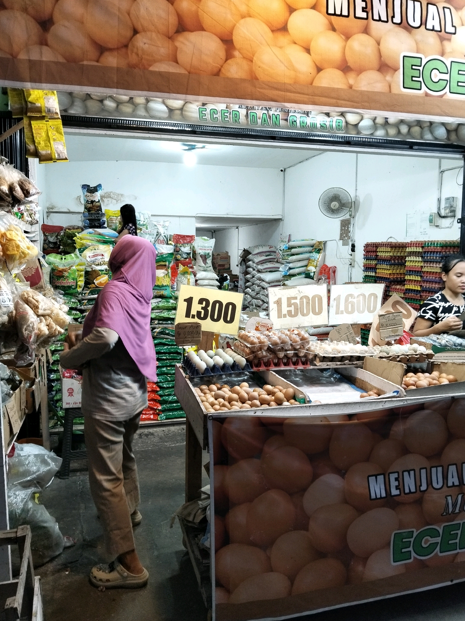 The image depicts a street-side market stall in Arang Limbung, Indonesia, specializing in the sale of eggs and other dry goods. The stall is covered with a banner featuring an advertisement for selling eggs. In the foreground, a woman wearing a pink hijab stands facing away from the camera, looking towards the stacked goods. She is wearing a beige outfit and light-colored slip-on shoes. To the right, another woman is visible behind a counter, wearing a black and white patterned shirt and looking towards the left.
Various types of goods are displayed for sale, including large quantities of rice sacks, packaged snacks, and numerous cartons of eggs in different sizes and colors. Prominently displayed on the stall are price signs. One sign reads "1.300" in large black numerals on a yellow background, and another shows "1.500" and "1.600" on white signs. The market appears to be outdoors, with a concrete floor. The overall atmosphere suggests a typical bustling local market scene.