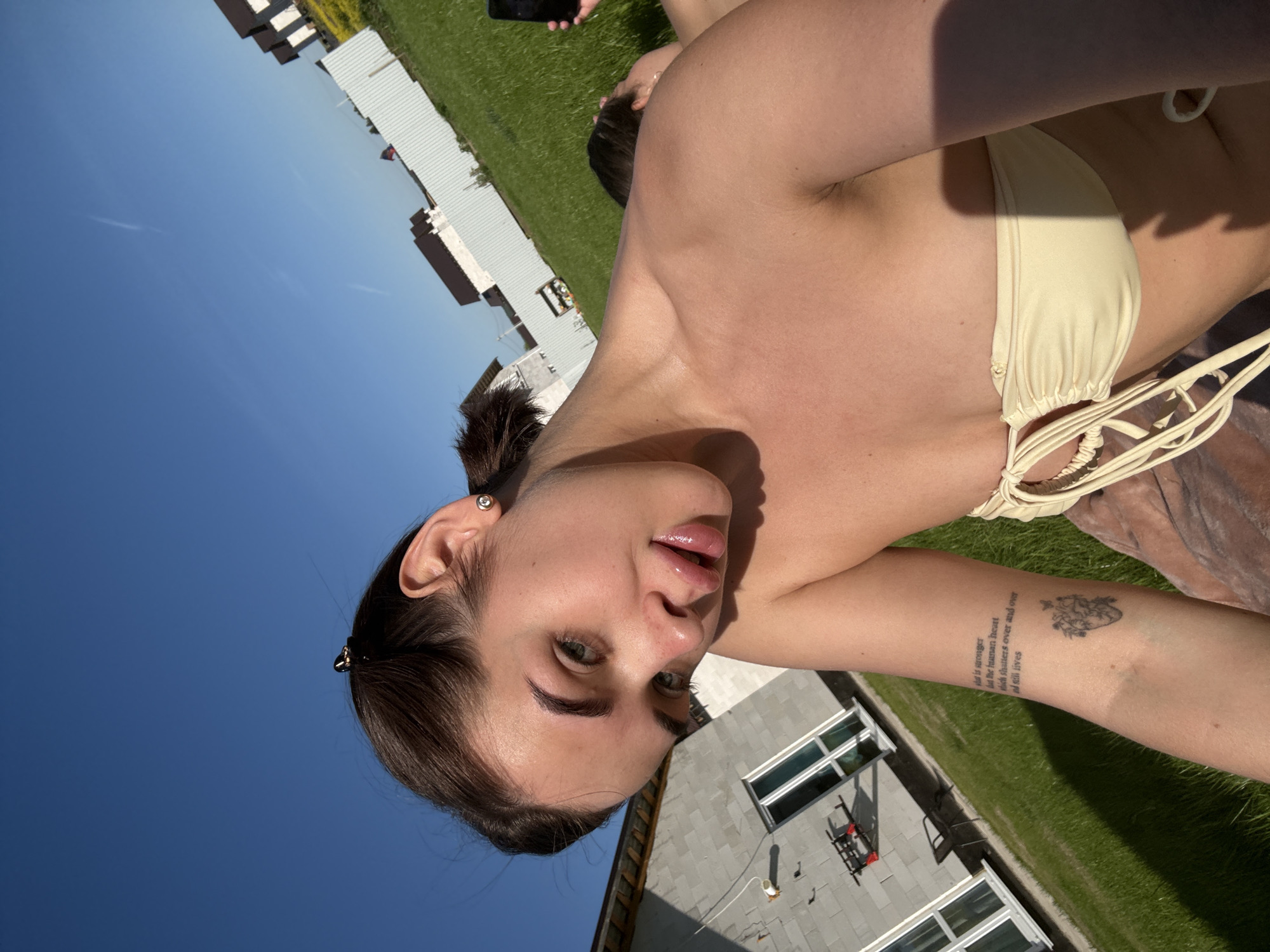 A woman is lying outdoors on a sunny day in what appears to be a residential area. She is wearing a light-colored bikini top. Her dark hair is pulled back, and she has a tattoo on her left forearm featuring text and a floral heart. She is positioned in front of a blue sky and a house with light-colored siding and several windows. Green grass surrounds the area. The photo's location might be in the vicinity of Лобаново, Russia, based on geographical context, but it can't be conclusively determined.