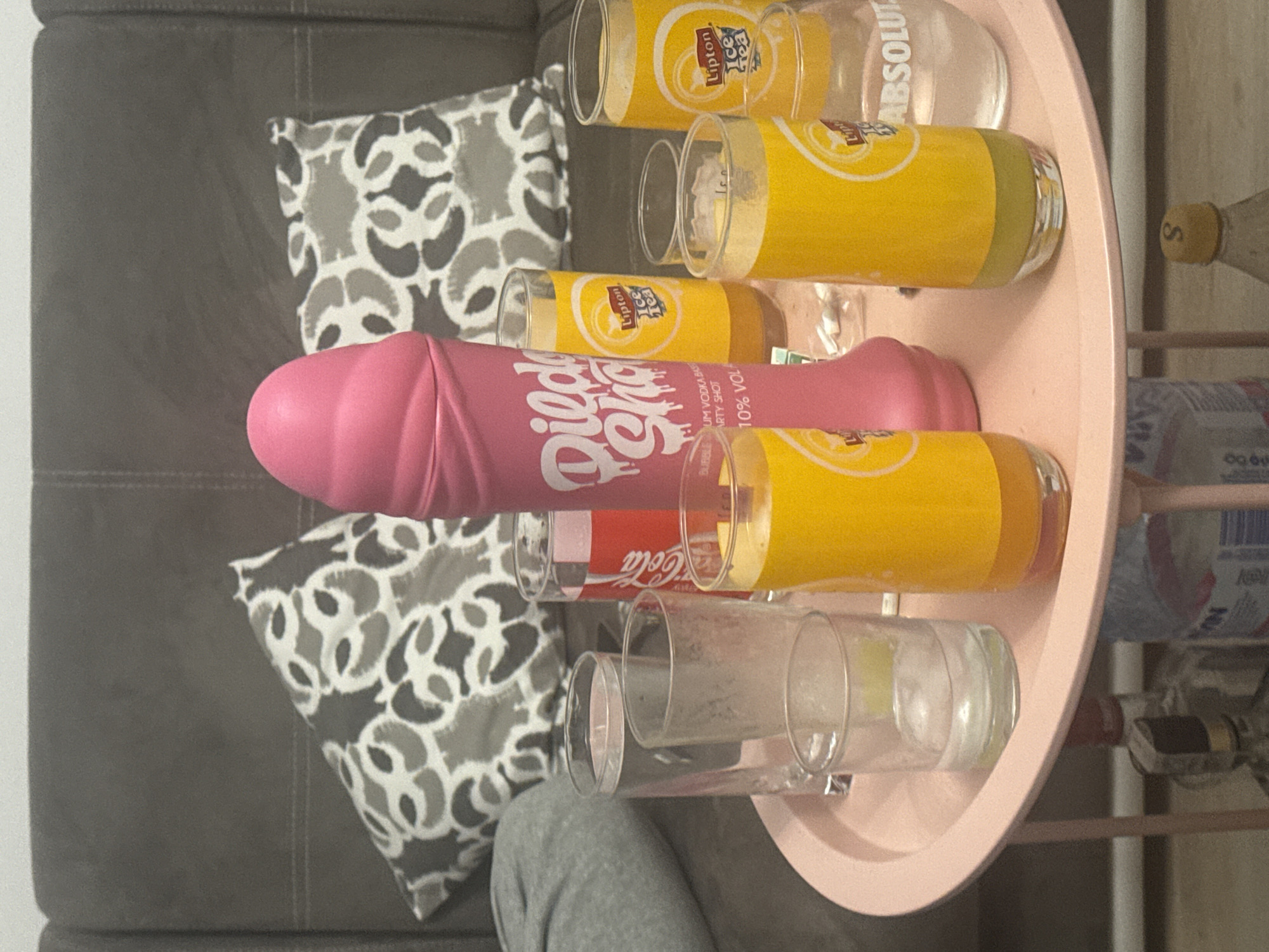 The image shows a pink, phallic-shaped party shot bottle with "Dildo Shot" written on it, along with a collection of glasses and beverage containers including Lipton Iced Tea, Coca-Cola, and Absolut Vodka, arranged on a light pink serving tray. The tray is situated near a grey sofa with a patterned pillow. Part of a person wearing grey clothing is also visible. The location is not specified in the image or OCR.