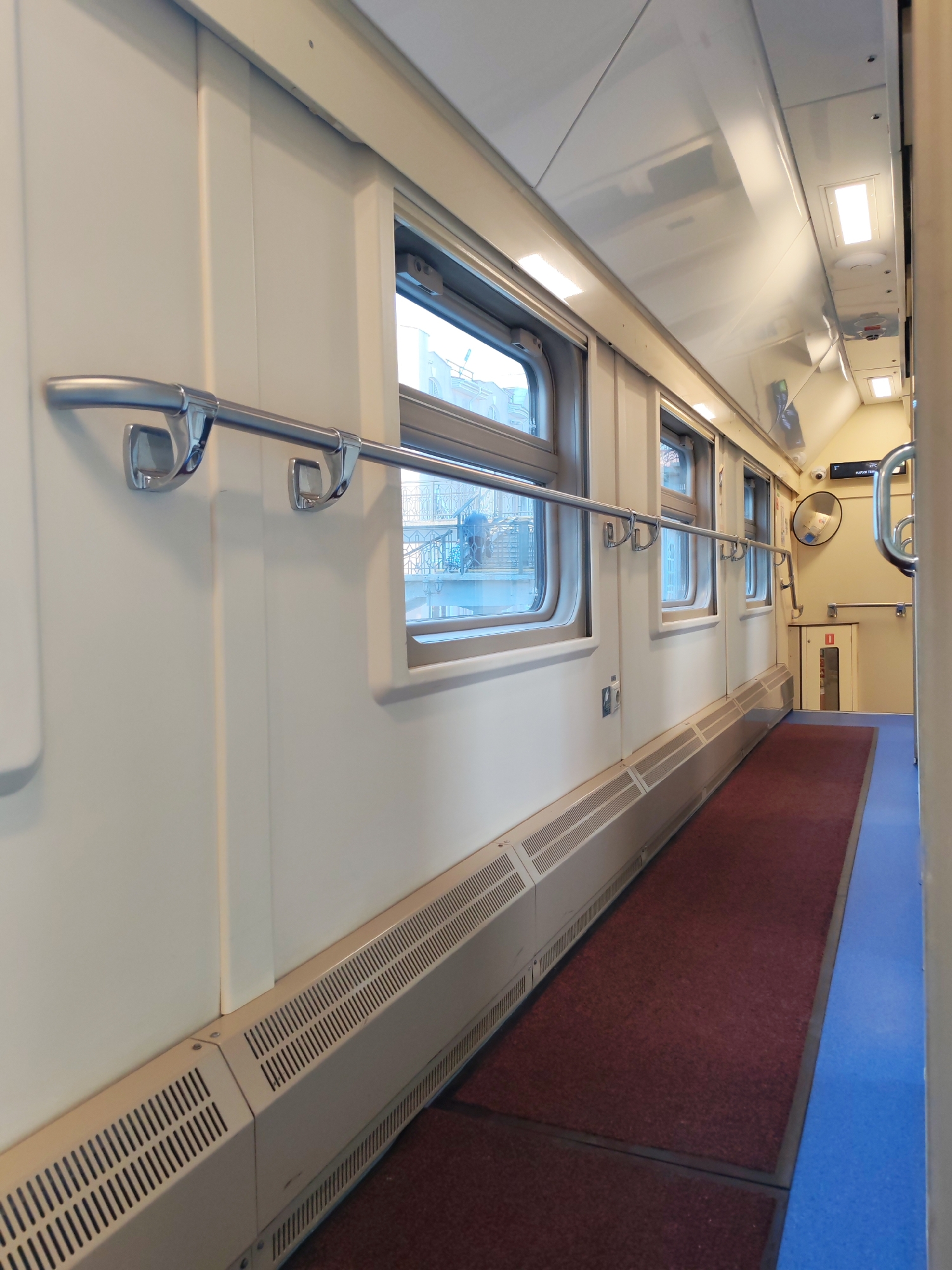 This media file presents a well-lit interior view of what appears to be a train or tram car's aisle. The perspective is looking down a long, narrow corridor, with windows lining one side. The dominant colors are off-white for the walls and ceiling, a deep red carpet runner on the floor, and a bright blue trim or flooring alongside the carpet.
Along the off-white wall with windows, a chrome handrail is prominently featured, secured by several U-shaped brackets. Below the windows, a long, low heating or ventilation unit with perforated grates runs the length of the corridor. The windows show glimpses of an urban or suburban environment outside, with buildings and what might be a bridge or overhead structure visible through the panes, suggesting it's daytime.
The ceiling is made of several light-colored panels, some of which feature rectangular light fixtures providing illumination. Towards the end of the visible corridor, there's an opening or doorway. On the right side of the corridor, partially obscured, is another wall. A convex mirror is mounted on this wall, and below it, a small cabinet or box with a red frame and a clear panel, possibly indicating emergency equipment like a fire extinguisher. Above the mirror, a small digital display or sign is visible, along with a security camera mounted high on the wall. Another chrome handrail is also present on this side near the end of the corridor.
No people are visible in the scene, and no specific activity is taking place, giving the impression of a temporarily empty space, perhaps between stops or at a depot.
Visible text:
On the small digital display above the convex mirror, some Cyrillic text is discernible. The top line shows "°С" (degrees Celsius), indicating a temperature reading. Below it, the word "НАРУЖН.ТЕМП" which translates to "OUTSIDE TEMP" (наружная температура). The displayed temperature value is not clearly legible.