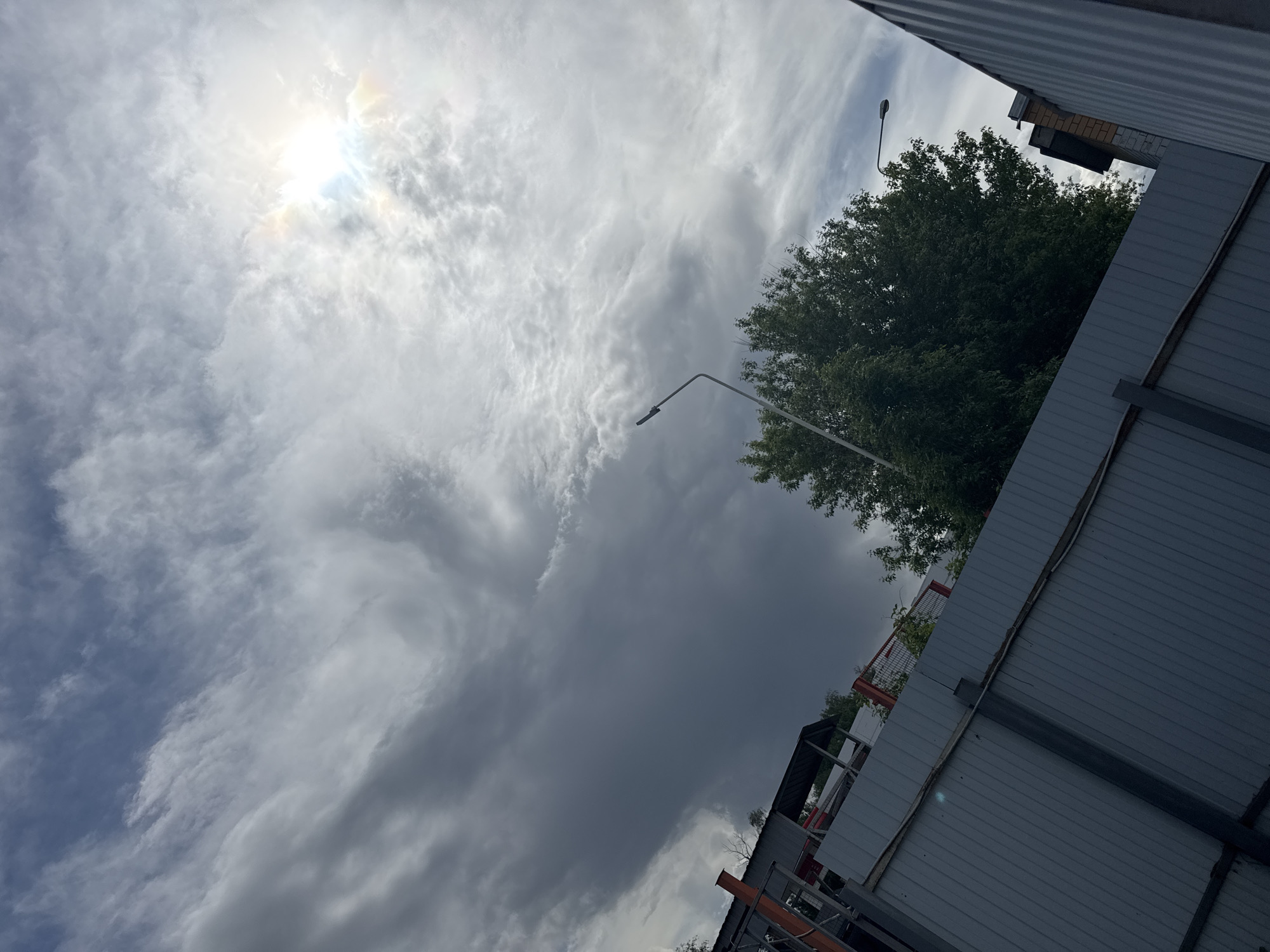 The image is taken from a low angle, looking up at a cloudy sky. The sun is visible through the clouds in the top left corner. A tree is in the right side of the frame, with a street lamp extending toward it. A corrugated metal fence and portions of buildings are on the right and bottom sides of the image. Based on visual cues and the user-provided location, the setting could plausibly be in Balashikha, Russia, but there are no specific elements confirming the city. No people are visible.