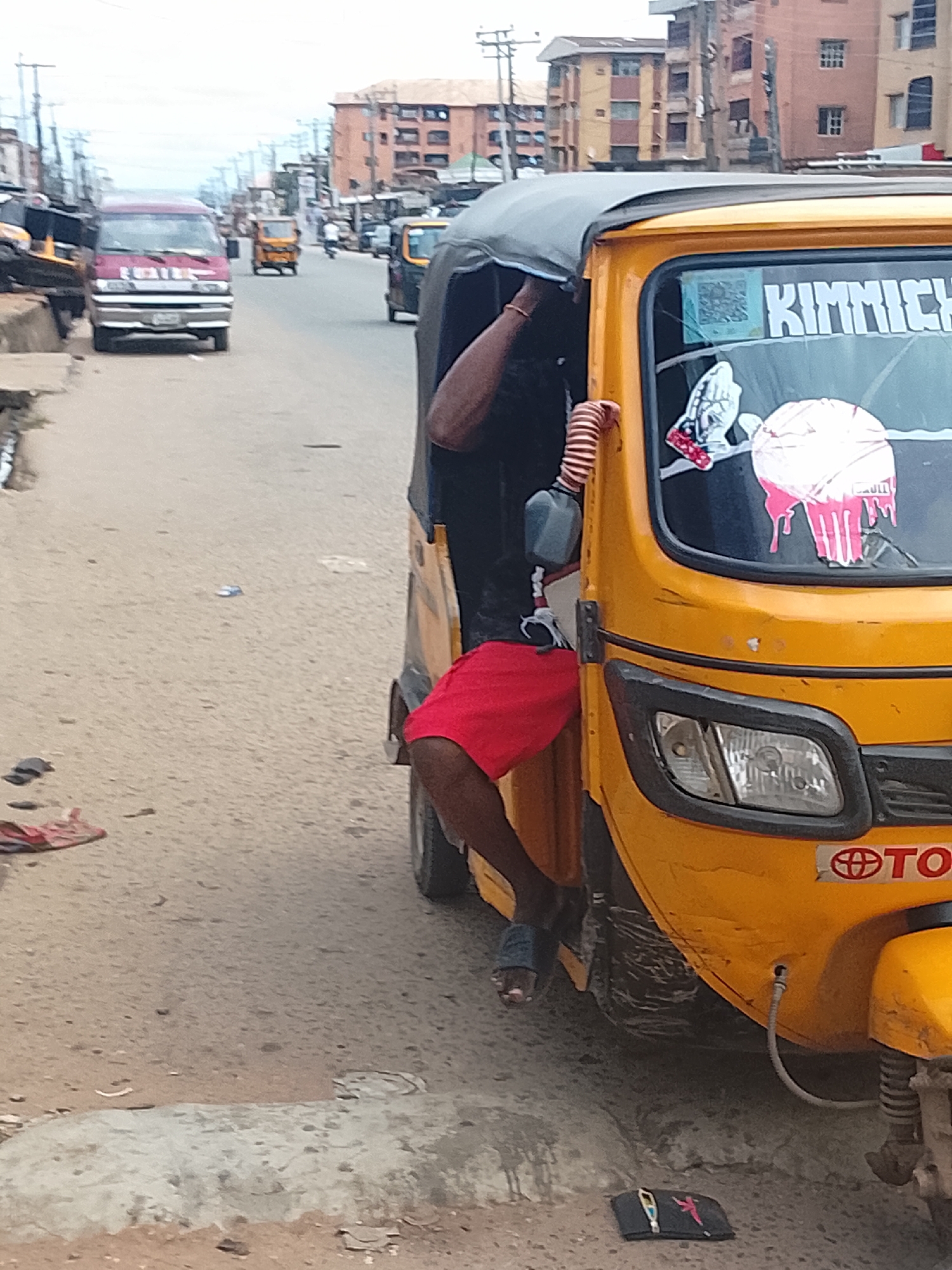 An outdoor, eye-level, medium shot shows a yellow auto-rickshaw parked on a dusty, paved road, with the front passenger side of the vehicle dominating the foreground. The auto-rickshaw has a black fabric roof and the front windshield displays stickers. One sticker reads "Kimmich", another is a QR code, and two other stickers are red and white illustrations; one appears to be praying hands, and the other is a pink dripping abstract shape. A person's lower body, clad in red shorts and dark blue sandals, is visible in the passenger seat. A black striped tube is attached to the side of the vehicle. The background shows a street scene with other vehicles, including a red minivan and several auto-rickshaws, as well as buildings lining the street. The sky is overcast. The road has some debris and a discarded black pouch with a key and a pink object on it.