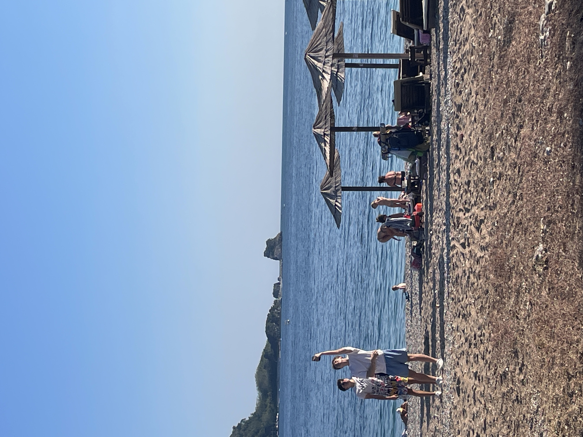 The image shows a beach scene in Budva, Montenegro, on a sunny day. The sky is clear and blue. The water is calm and blue. There are people on the beach, some sunbathing under umbrellas and others standing near the water's edge. In the foreground, two people are taking a selfie with a mobile phone. The beach is sandy and has some pebbles. In the background, there are some islands and green vegetation. There are also umbrellas and chairs available for beachgoers.