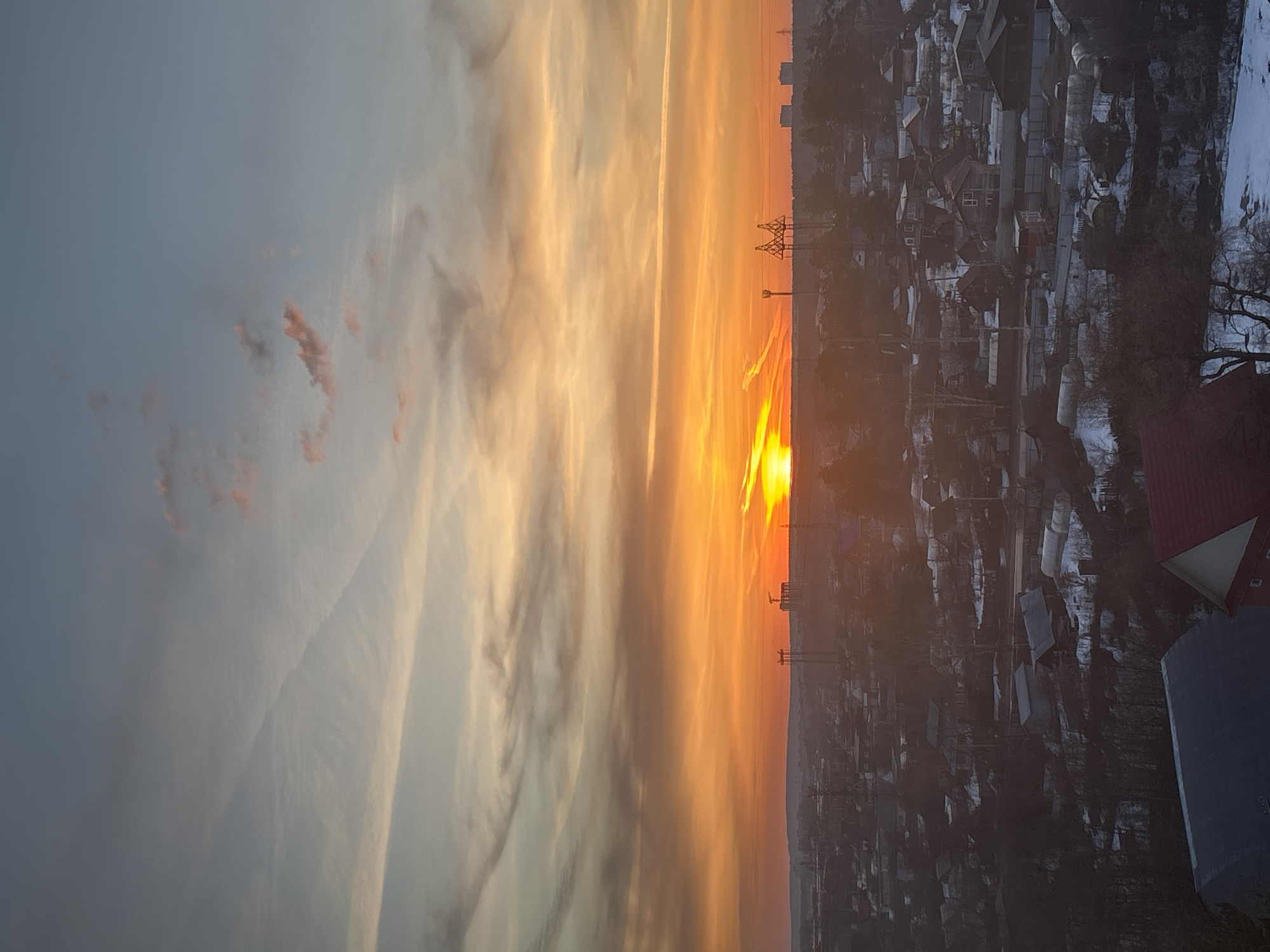 The image shows a sunset view over a residential area, possibly in Chaikovsky, Russia. The sun is setting on the horizon, radiating orange and yellow light across the sky and illuminating the clouds. To the right of the sunset, there is a cityscape with houses and buildings, some of which appear to be covered in snow. There are also power lines and trees visible in the landscape. The image is vertically oriented, presenting a wide perspective of the scene.