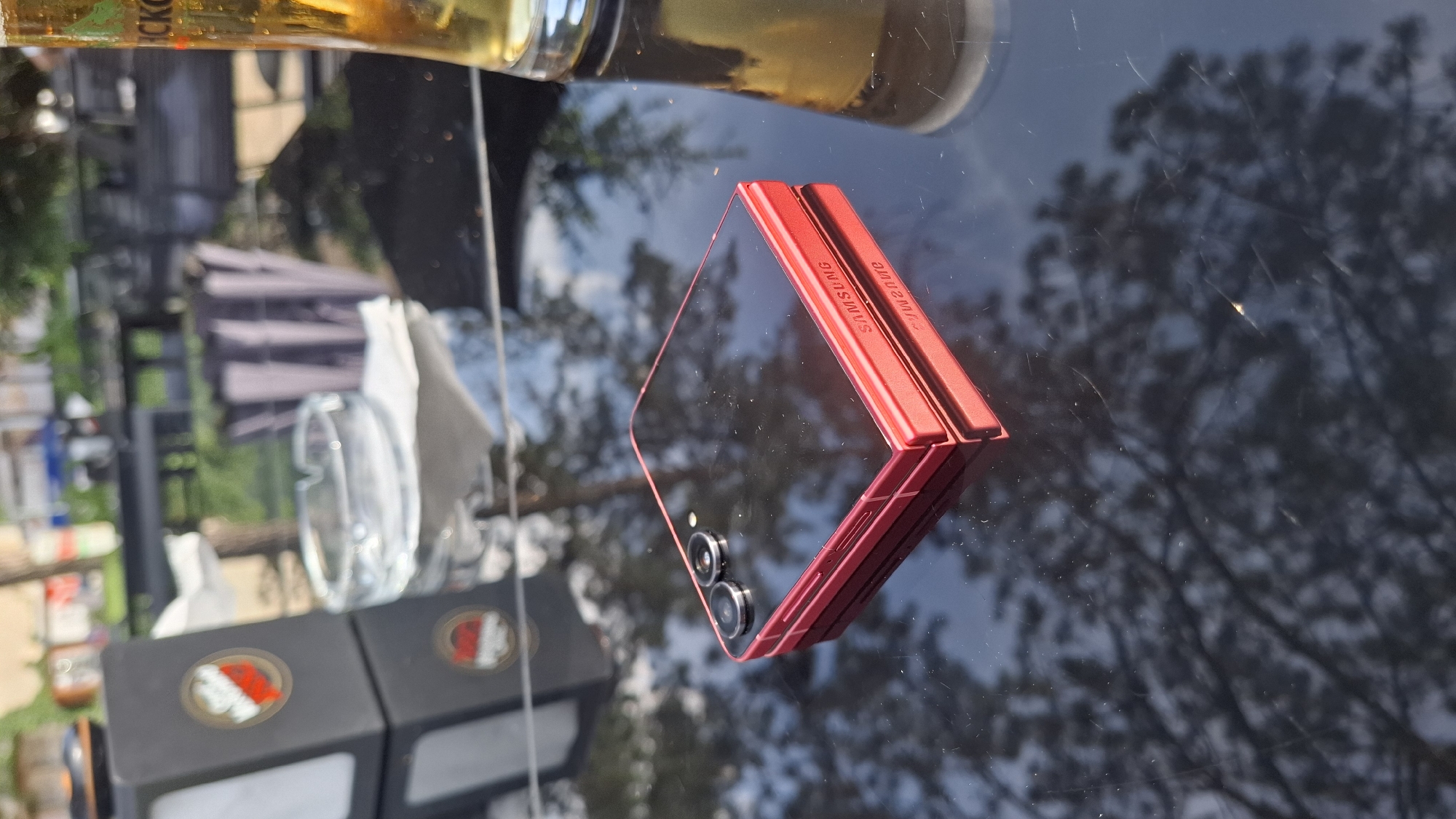 A red Samsung flip phone is resting on a reflective black table. The phone is closed and positioned at an angle, with its screen facing upwards. The brand name "SAMSUNG" is visible in white text on the side of the phone. To the left of the phone, a black napkin dispenser with a circular red and white logo is partially visible. An ashtray sits behind the napkin dispenser. To the right of the phone, a glass of amber-colored liquid, likely beer, is partially visible, with a portion of its label showing green and white text. The table's surface reflects the surrounding environment, showing blurred trees and outdoor seating arrangements, suggesting an outdoor setting such as a patio or beer garden. The lighting indicates it is daytime.
