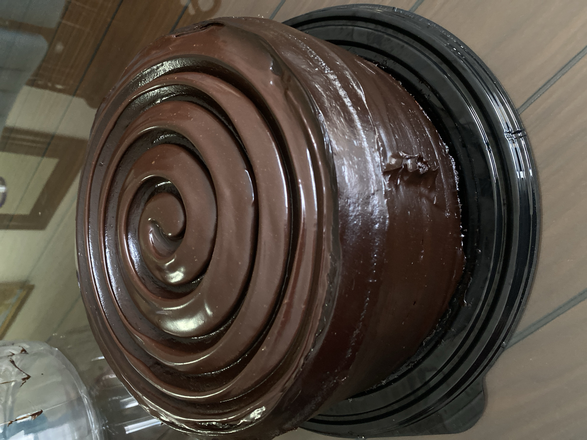 A chocolate cake sits on a black plastic dish on a brown wooden surface. The cake is round with a spiral of chocolate frosting on top. The cake and dish are sitting on a table indoors.
