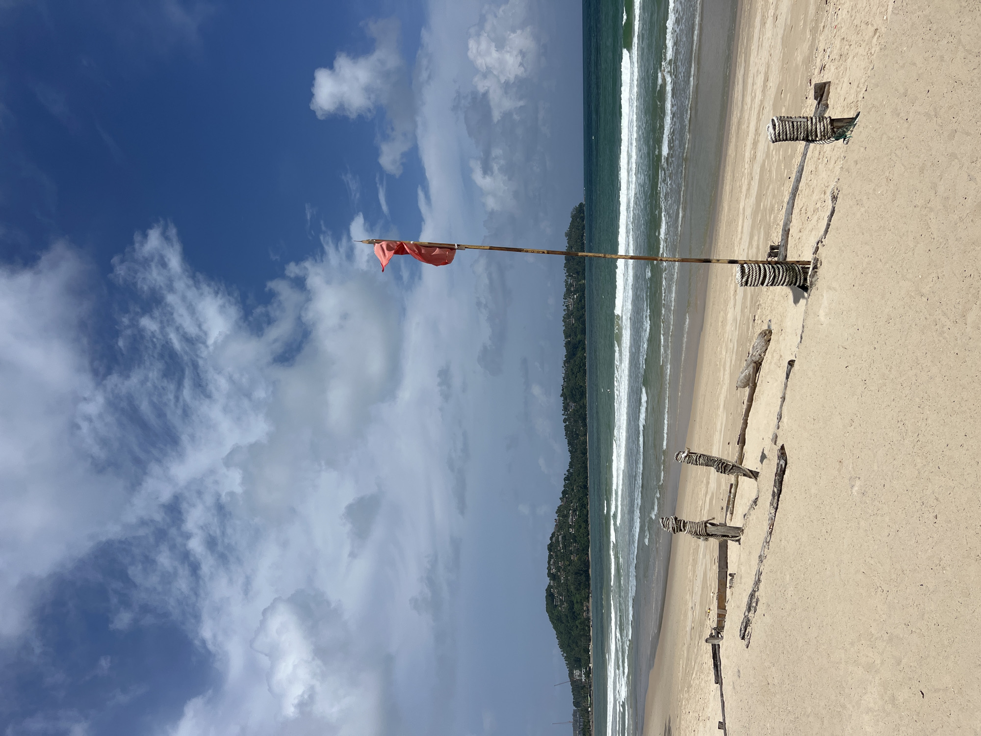 A bright, sunny day at a beach in Choeng Thale, Thailand, is depicted. The foreground is a sandy beach with some weathered wooden posts and debris. A tall wooden pole with two red flags stands prominently in the center of the frame, indicating potentially hazardous conditions in the ocean. The middle ground shows gentle waves rolling onto the shore, with the turquoise ocean extending to the horizon. In the background, a lush green hill dotted with buildings overlooks the sea, suggesting a populated coastal area. The sky above is a vibrant blue, with scattered white clouds adding texture to the scene. The overall atmosphere is serene and idyllic, typical of a tropical beach setting.