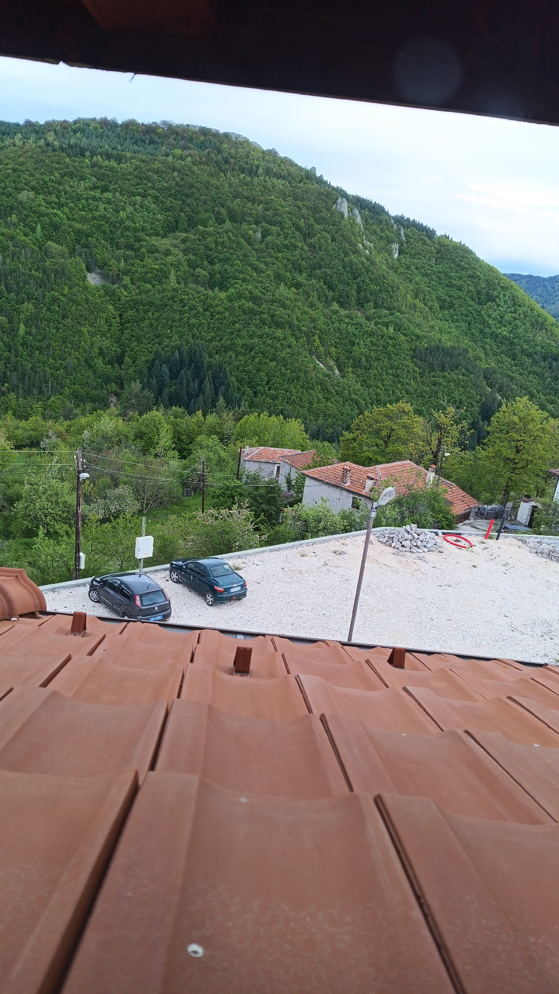 The image presents a scene in Vrata, Bulgaria, viewed from a high vantage point on a red-tiled roof. The scene includes two cars parked on a gravel surface adjacent to several buildings with red-tiled roofs. A wooden beam is in the foreground with a clear view of a densely wooded hill. A utility pole is visible near the cars, and the sky is overcast.