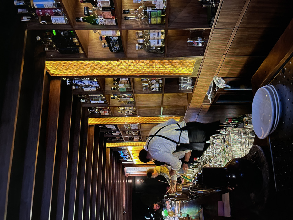A bar interior features dark wood walls and shelves stocked with liquor bottles, illuminated by warm lighting. A bartender in a white shirt and black apron is working behind the bar, tending to glassware and drinks. Other staff members are visible in the dimly lit background. Plates are stacked at the edge of the bar.
