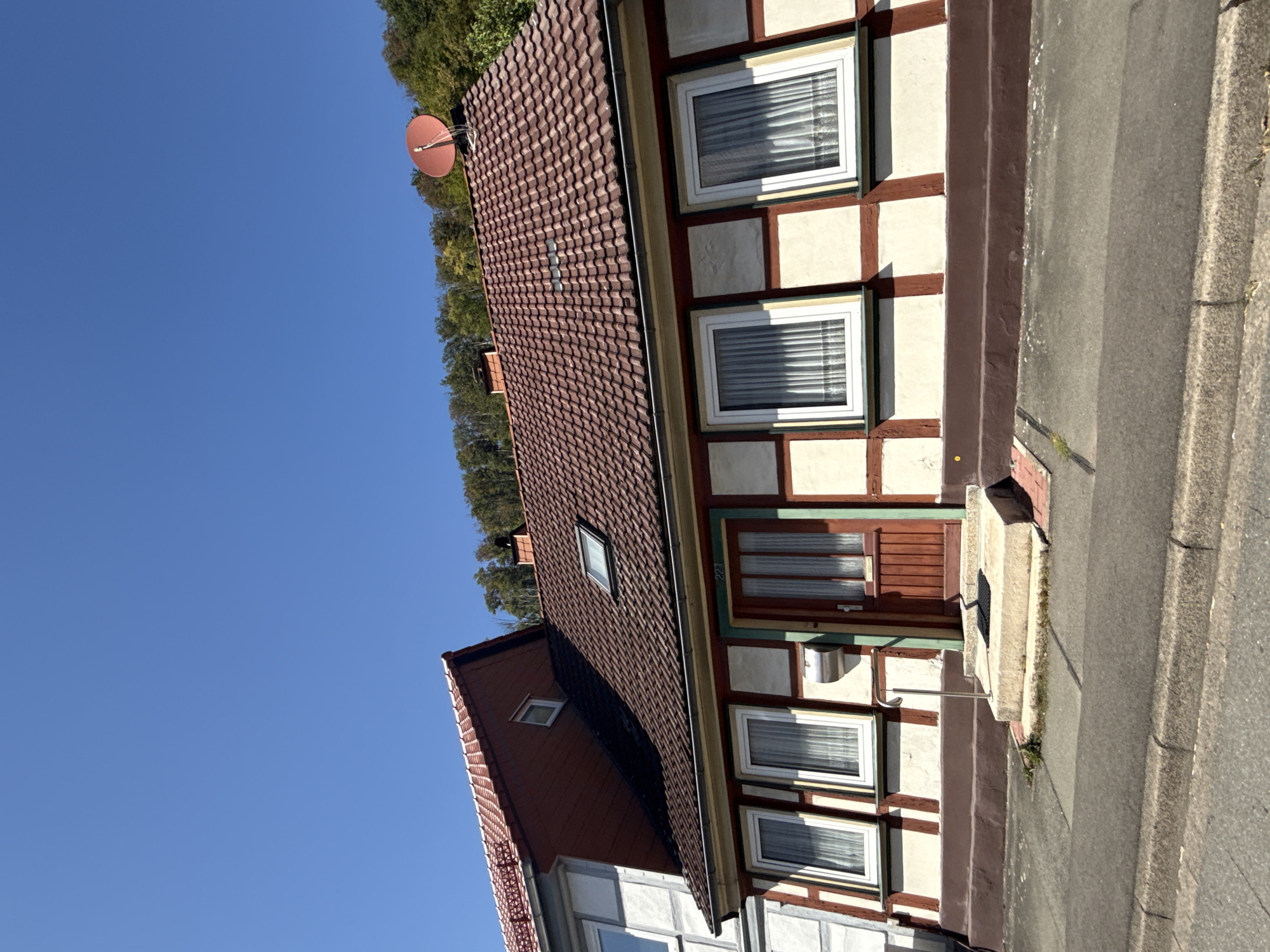The image shows a side view of a two-story half-timbered house under a clear blue sky. The house has a red tiled roof with a dormer window. The facade is cream-colored with dark brown timber framing. There are several white-framed windows with white curtains. A wooden door with glass panes is visible on the right side of the facade, and a metal mailbox is mounted next to it. A small set of concrete stairs leads to the entrance. The house is situated on a street with a paved road and sidewalk. Trees and a hillside are visible in the background. The house is located at Kirchberg 221 in Bad Lauterberg im Harz, Germany.