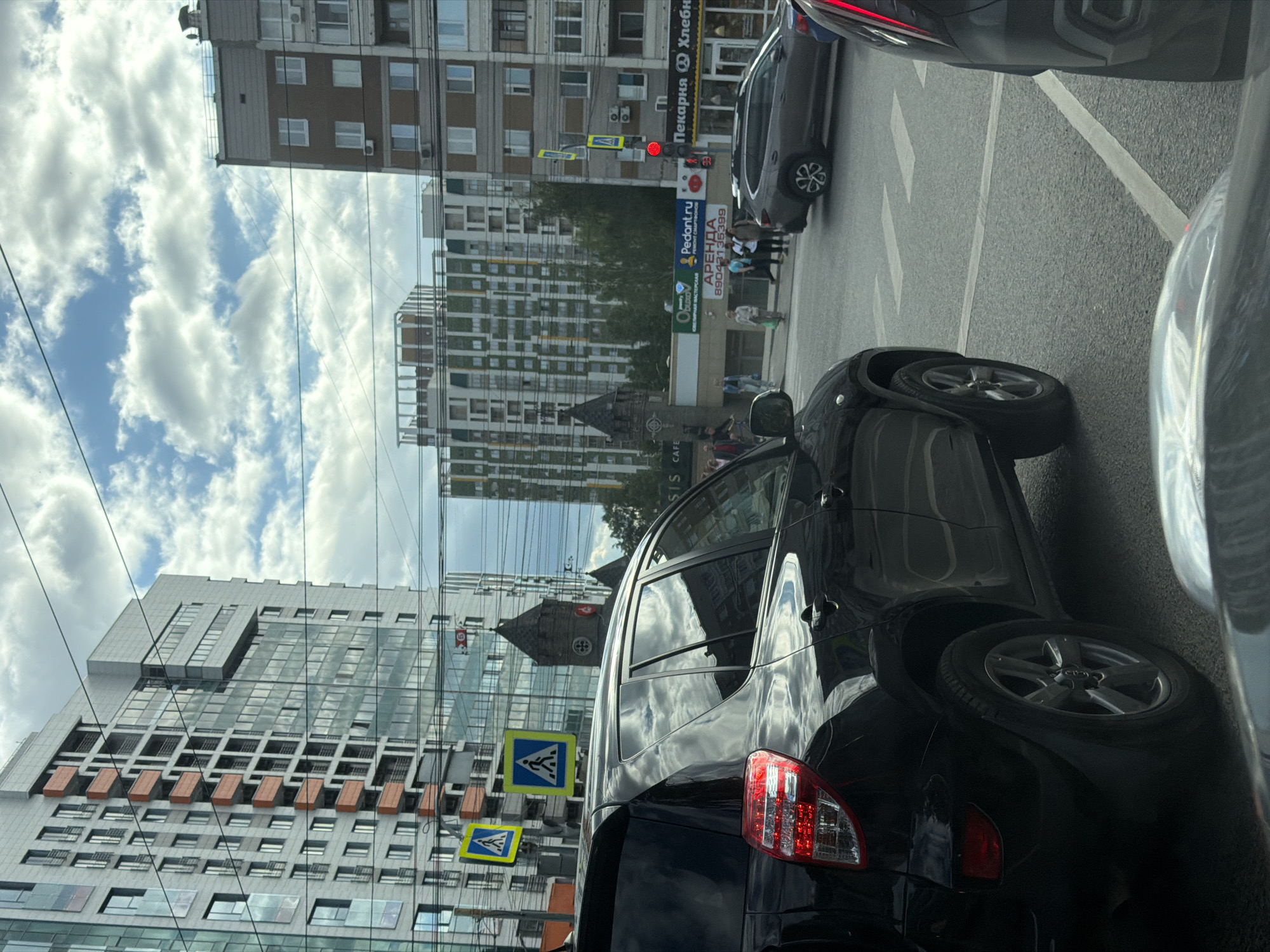 The image shows a street scene in Izhevsk, Russia, with several cars and modern buildings. In the foreground, a black SUV is stopped at a traffic light, likely at an intersection. To the left, there are pedestrian crossing signs. The background features several tall, contemporary buildings, some with extensive glass facades, and a more traditional-looking building with a dark, pointed turret. The sky is partly cloudy. Pedestrians are visible on the sidewalk and crossing the street. A red traffic light is illuminated. Signs for businesses, including "Pedant.ru" and "Пекарня" (Bakery), are visible on the buildings.