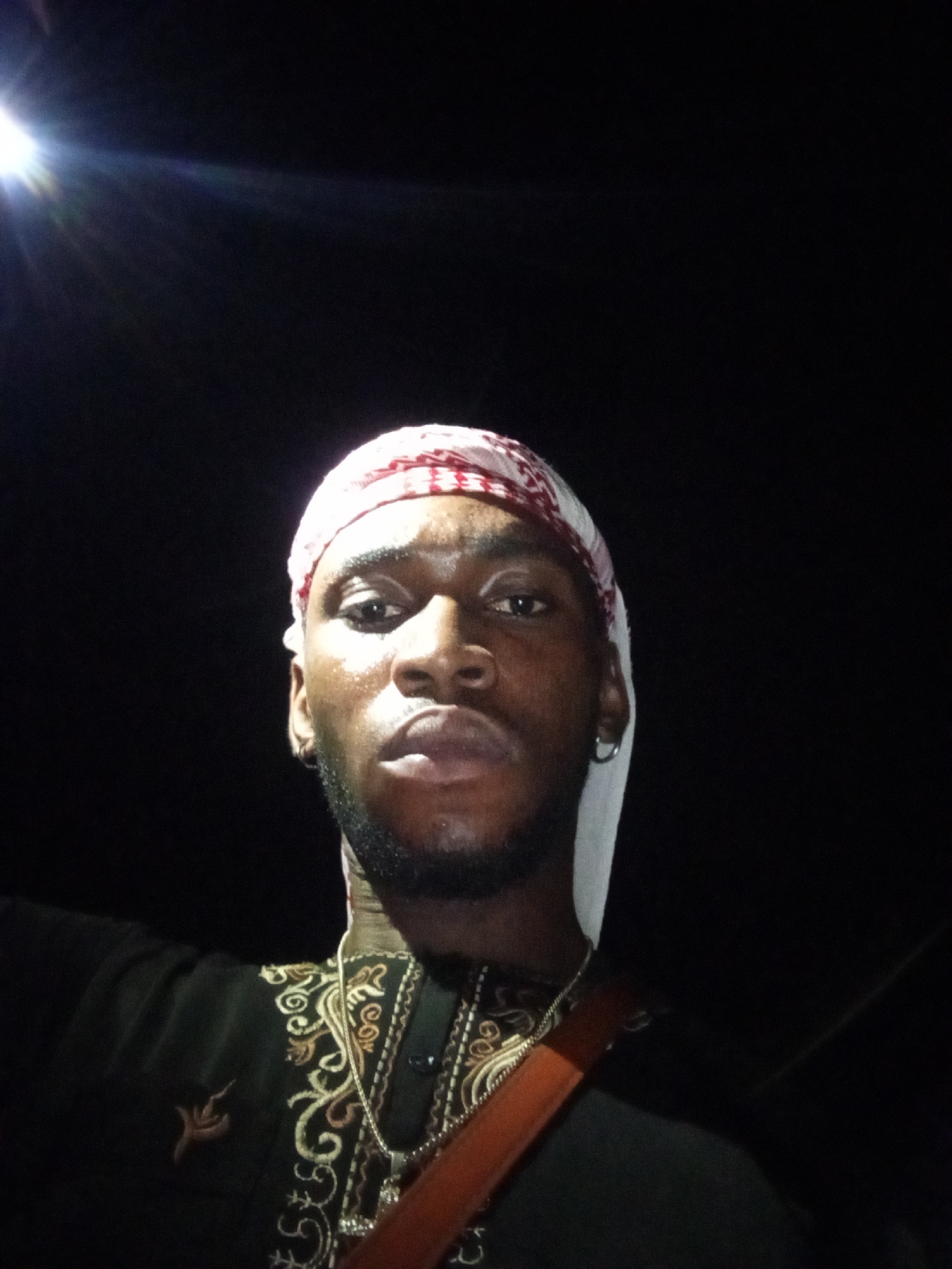 A man is standing outside at night. He is wearing a head covering with a red and white pattern. He also has on a dark shirt with gold-colored embroidery. He is wearing a silver chain around his neck. The environment is dark, with a bright light source illuminating the scene from above. I have no information about the city.