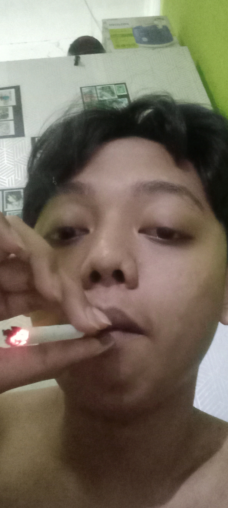 A close-up selfie shows a person's face and upper chest. The person has dark hair and is holding a lit cigarette to their lips with their right hand. The background appears to be an indoor setting with a wall and some framed pictures. The image is taken from a low angle, focusing on the individual's facial features and the act of smoking. The specific location in Argasoka, Indonesia, is not discernible from the visual information provided.