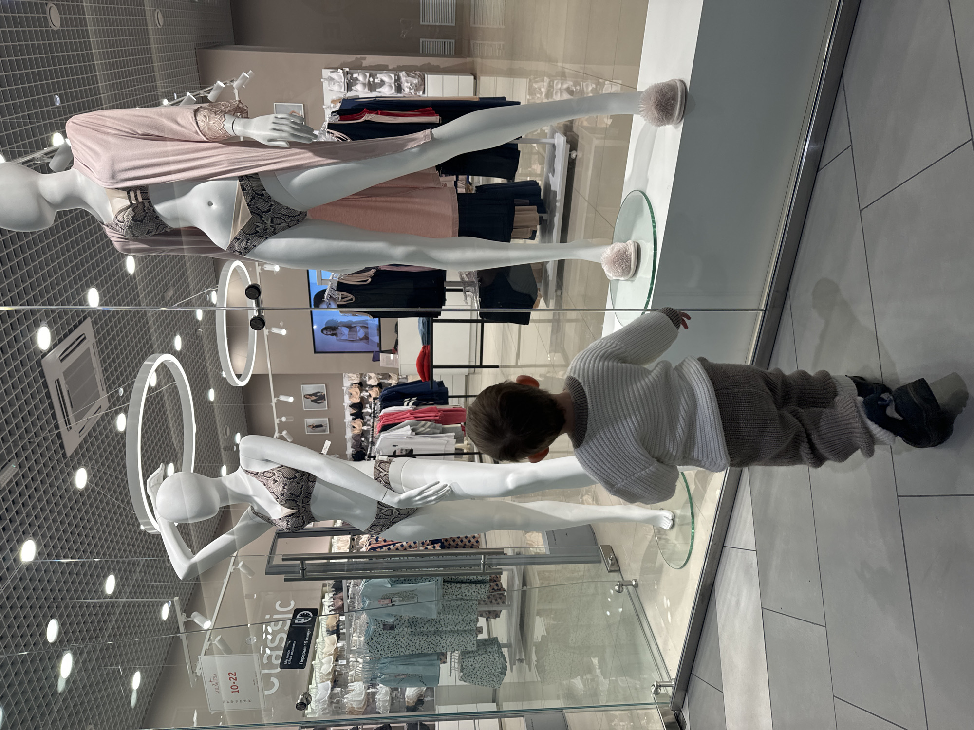A child is observed examining a display of mannequins in a shop window at an unknown retail location in the city of Kriukovo, Russia. The mannequins, styled in undergarments, are positioned within the shop window. The child, dressed in casual attire, is peering into the display. The shop window is made of clear glass.