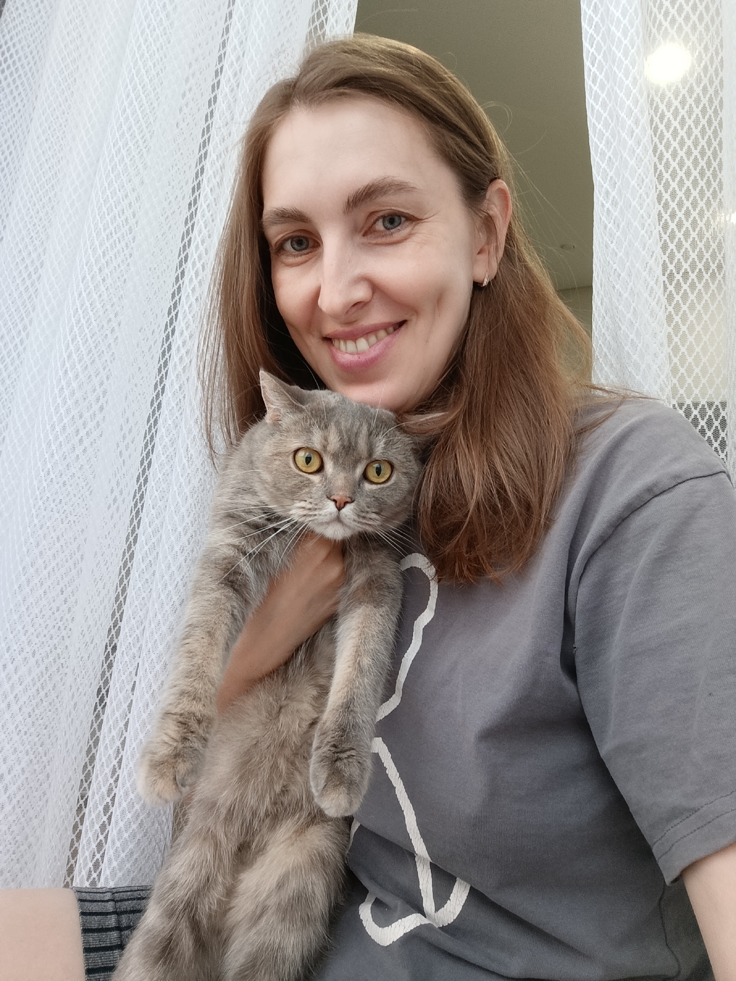 In a close-up, selfie-style shot, a woman with light brown hair and blue eyes smiles at the camera. She is holding a grey and white tabby cat with bright yellow eyes. The woman is wearing a grey t-shirt with a white line drawing of a cat on it and a striped skirt. She is positioned in front of a white, patterned sheer curtain. The lighting suggests an indoor setting.