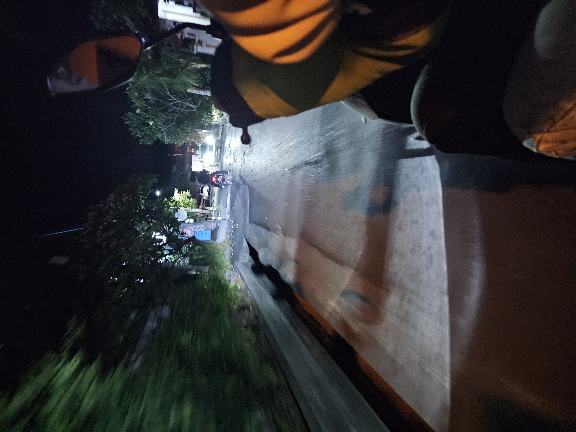 The image captures a first-person perspective of a motorcycle ride at night on a road in Denpasar, Indonesia. The road appears wet, reflecting the light from a car's headlights, which are visible in the distance. A person on another motorcycle is also ahead, illuminated by their taillight. Lush green trees line the left side of the road, and a stone wall runs alongside it. The sky is dark, indicating it's nighttime. The rider's hand, covered by a jacket with green and yellow stripes, is visible on the right side of the frame, gripping the handlebar. The motion of the motorcycle is evident from the blurred surroundings, suggesting a moving vehicle.