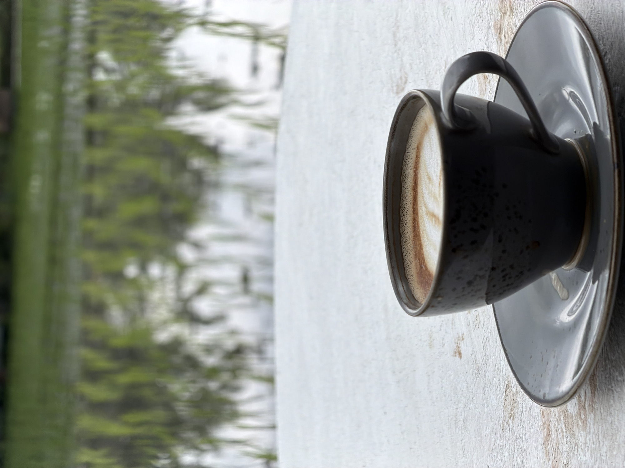 A dark gray mug of coffee with latte art sits on a gray saucer on a white wooden table. In the background, out of focus, is a green rice paddy with reflections of the sky and greenery in the water. This scene is located in Mengwi, Indonesia.