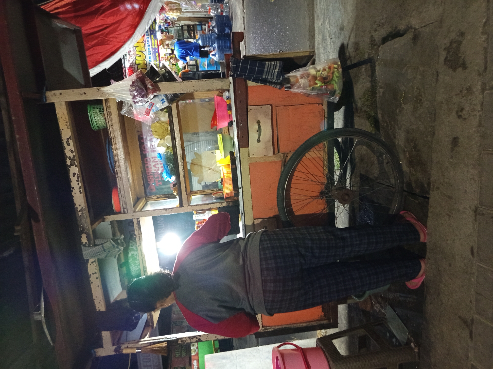 A person stands behind an orange food cart at night in Tebel, Indonesia. The cart has a wheeled base and a shelf display featuring produce in bags and bowls. The person, facing away from the camera, wears a grey and red long-sleeved shirt and plaid pants. A red plastic stool is visible to the left of the cart, and a red bucket rests on it. The ground appears to be paved with concrete and cobblestones. In the background, a few individuals are visible near a store.