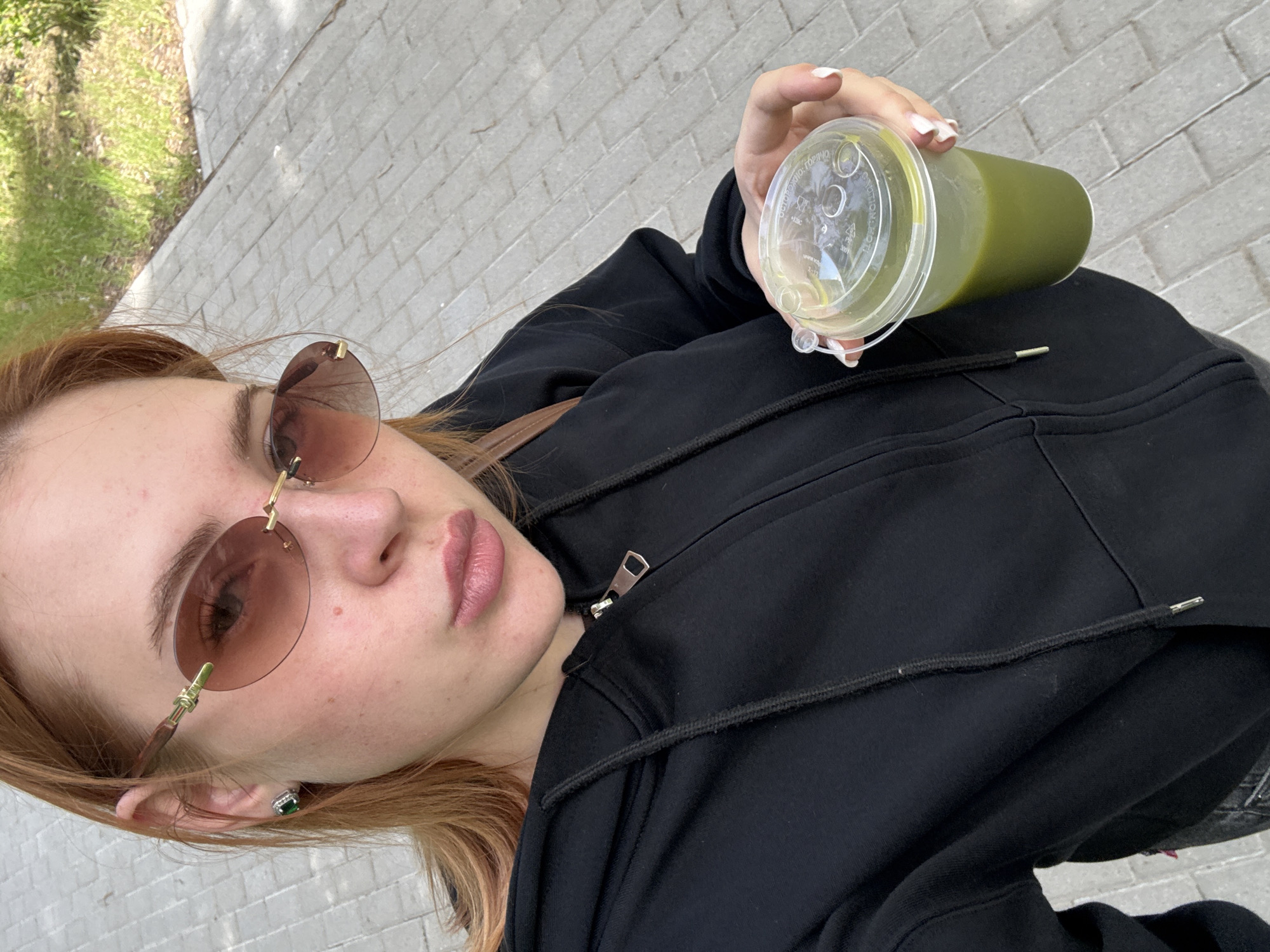 An angled, close-up selfie captures a young woman outdoors in Chaikovsky, Russia. She is looking directly at the camera with a neutral expression. Her light brown hair frames her face, and she wears rimless sunglasses with brown-tinted lenses and gold-colored details. A small earring with a prominent green stone and silver/white border is visible on her right ear.
She is dressed in a black zipped hoodie. Her right hand, with white-manicured nails, holds a clear plastic cup containing a bright green liquid, likely a smoothie or juice. Partially visible text on the cup's lid includes "ОСТОРОЖНО ГОРЯЧО" (Caution Hot) and "2-PP".
The background features a light-colored paved surface, appearing to be bricks or pavers, suggesting a sidewalk or outdoor area. To the left, blurred green grass and foliage indicate an outdoor, possibly park or urban setting. The scene is brightly lit, consistent with daytime.