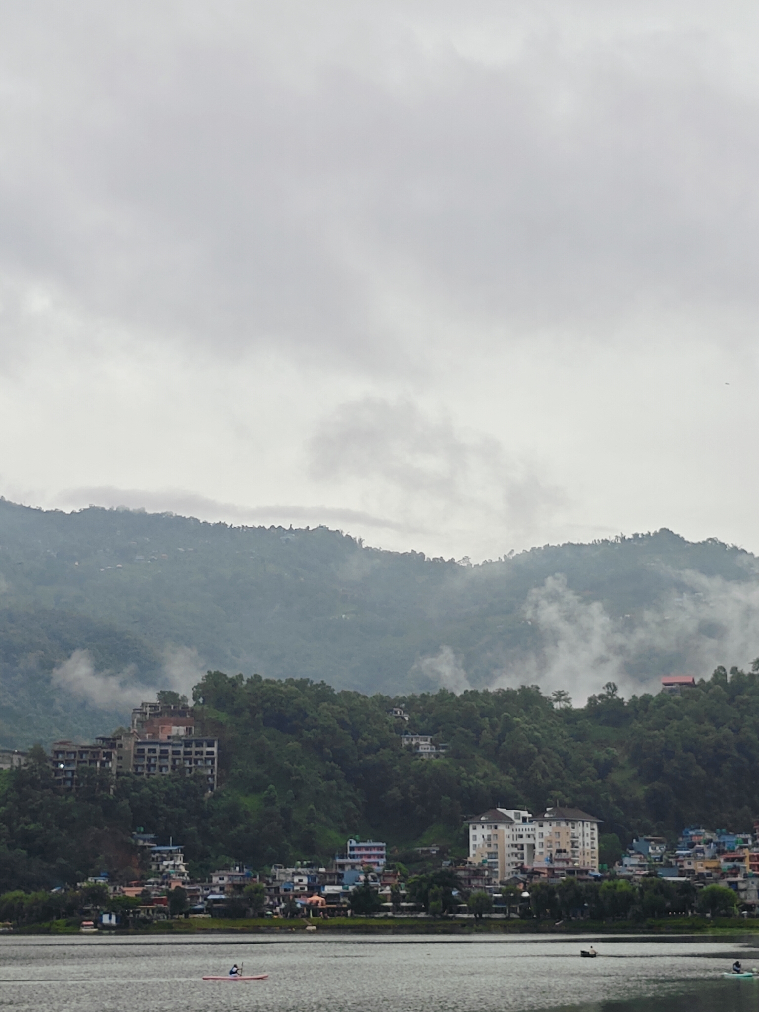 This image depicts the Lakeside area of Pokhara, Nepal, on a cloudy day. The foreground features a body of water, likely Phewa Lake, with a single person on a red paddleboard in the middle of the frame. In the midground, a shoreline is lined with a dense collection of buildings, ranging from multi-story structures with balconies to smaller, colorful houses. The buildings are nestled amongst green trees and vegetation.
Behind the town, a large, verdant hill rises steeply, covered in dense foliage. Several buildings, including a prominent multi-story structure with numerous windows and a more modern apartment building with light-colored facades, are visible on the hillside. Further up the hill, smaller houses with red roofs can be seen. The top of the hill is shrouded in mist and clouds, creating a layered effect and obscuring the distant mountain peaks. The sky is overcast with heavy, gray clouds, contributing to a muted and atmospheric scene.