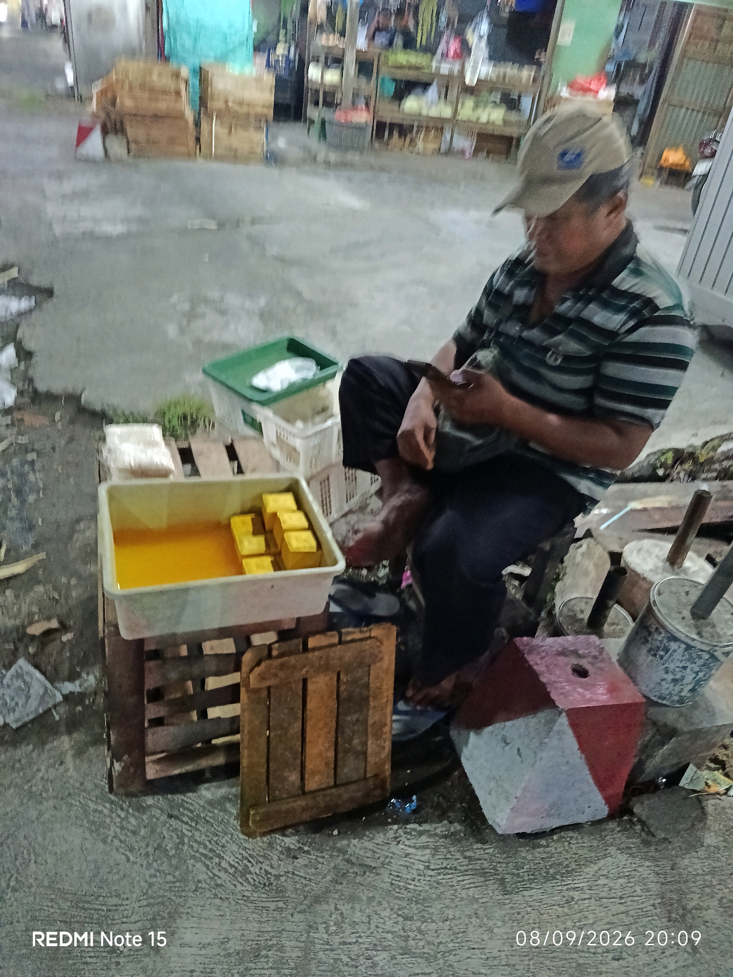 The image captures a street scene in Keboansikep, Indonesia, likely at dusk or night, given the ambient lighting. A person, appearing to be of Asian descent and wearing a striped shirt and a cap, is seated on a wooden pallet. They are holding a mobile phone in their hands and appear to be looking at it. In front of them is a large, light-colored rectangular container filled with a yellow liquid, with several cubed yellow items, possibly food products like tofu, submerged in it. To the left of the container, there are stacked packages wrapped in white plastic. Behind the seated person, there are wooden crates and shelves filled with various items, suggesting a market or vendor stall. To the right, a red and gray hexagonal concrete object with a hole in the top is visible, along with metal buckets. The ground is paved with concrete. The timestamp "08/09/2026 20:09" and "REDMI Note 15" are visible in the bottom corners of the image.