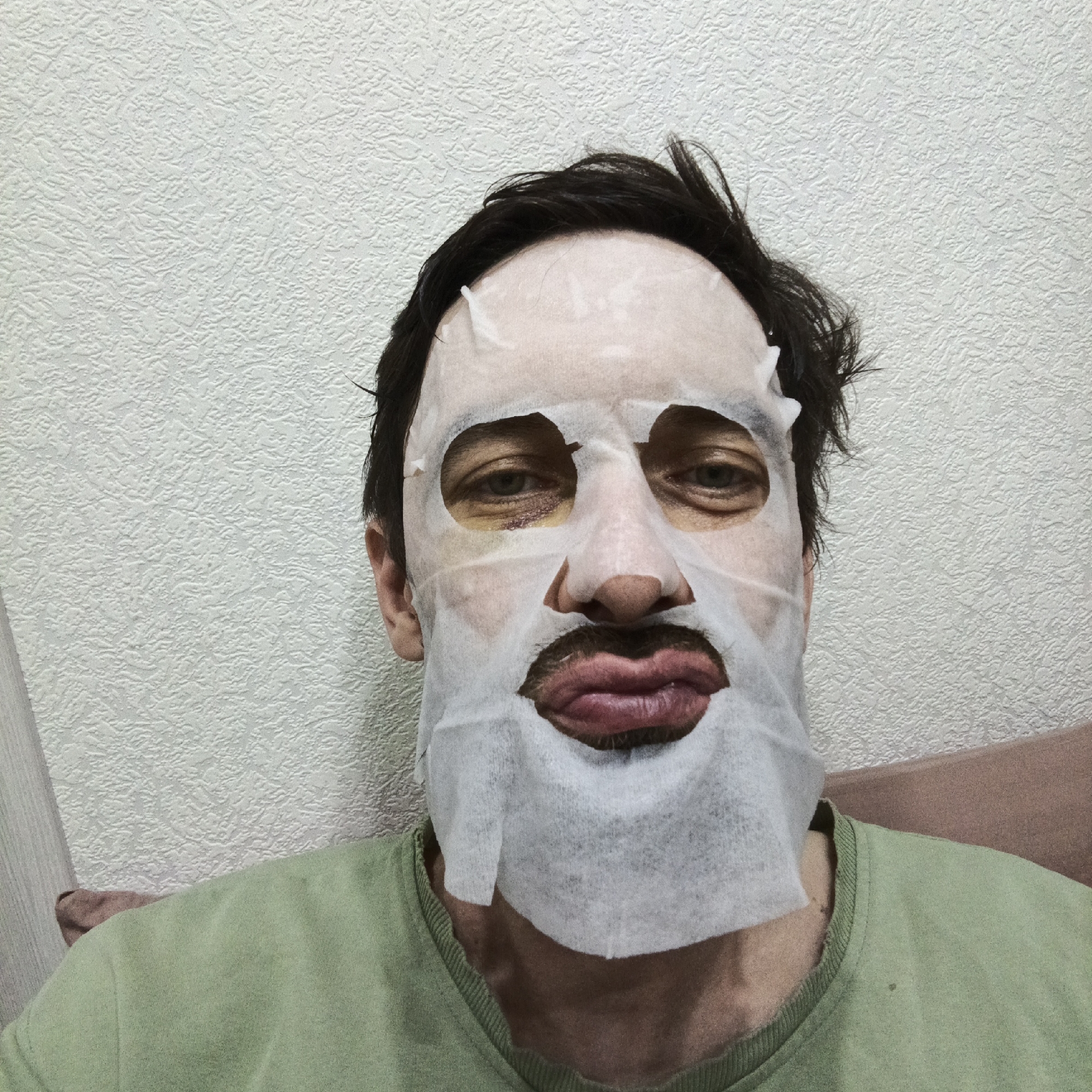A medium shot depicts a man wearing a white facial sheet mask with cutouts for the eyes, nose, and mouth. The man has dark hair and a green shirt. He is positioned indoors against a textured, light-colored wall, with a partially visible brown pillow or cushion behind him on the right side of the frame. The photograph was taken at an indoor location at Марковский, Russia.