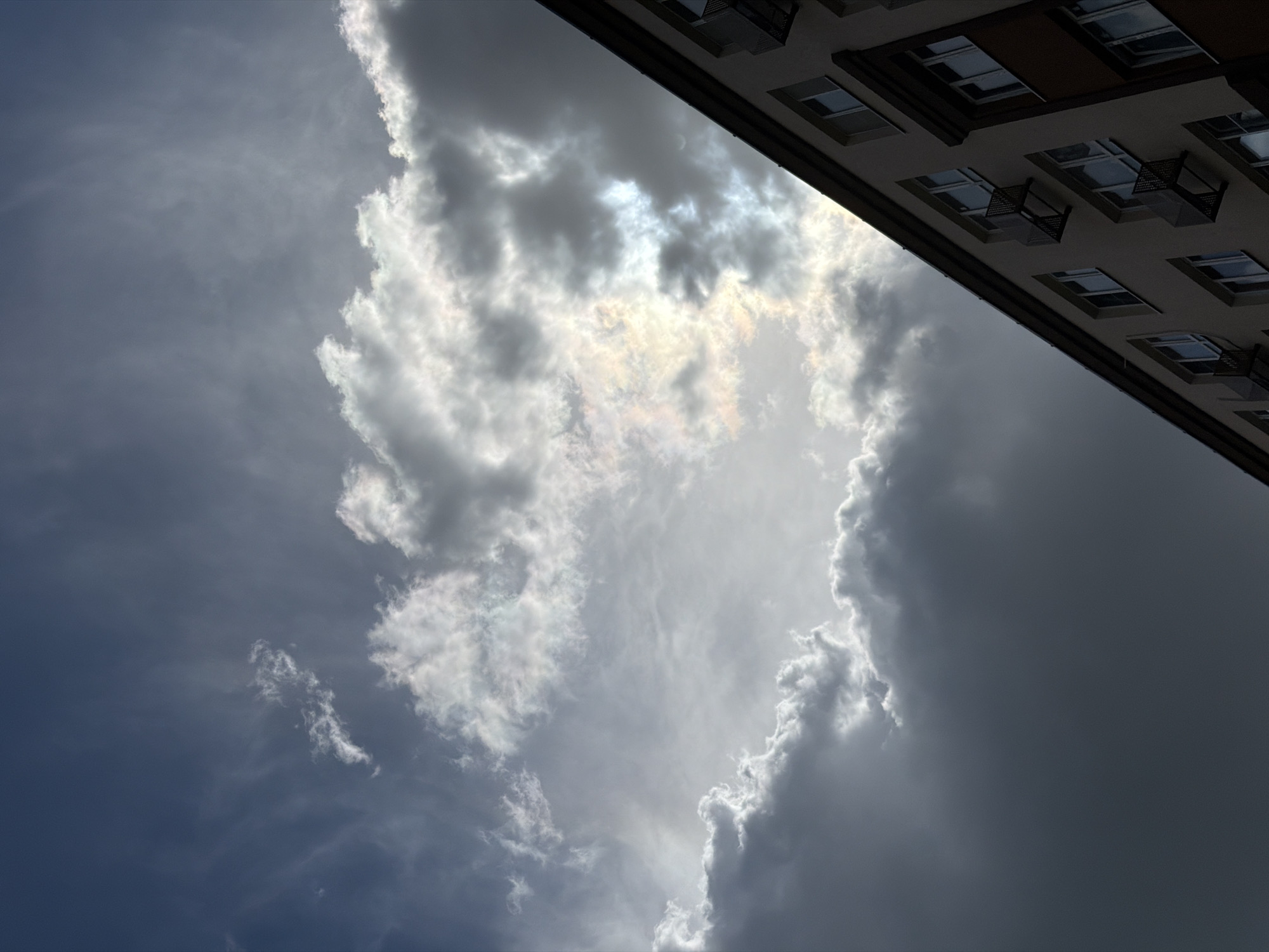 The image depicts a view of a cloudy sky with a building in the foreground, taken from the city of Lopatino, Russia. The sky is dominated by a mix of dark gray and white clouds, with patches of sunlight breaking through, creating a dramatic effect. The building, which appears to be an apartment complex, is seen from a low angle, its facade stretching diagonally across the lower right portion of the frame. Several windows, some with small balconies or air conditioner units attached, are visible on the building. The overall atmosphere suggests a potentially stormy or overcast day.