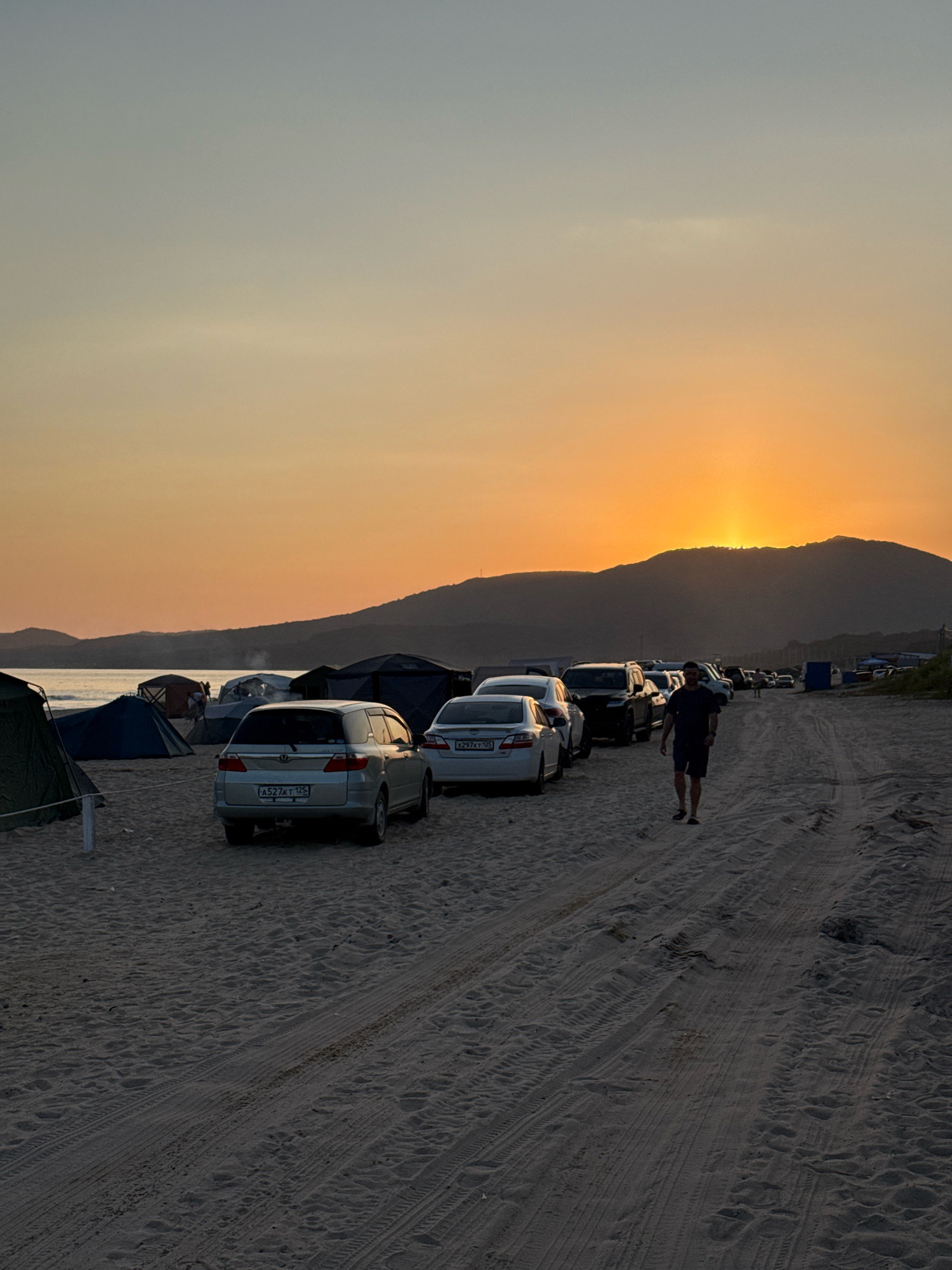 The image depicts a sunset scene on a sandy beach, likely located near the city of Anna, Russia, given the presence of a vehicle with Russian license plates. The sun is setting behind a silhouetted mountain range, casting a warm orange glow across the sky. Several cars are parked on the beach, some with their headlights on. Tents are set up near the waterline, suggesting a campsite. A single person is walking away from the camera on the sandy track, silhouetted against the setting sun. The tide is out, exposing a wide expanse of sand with visible tire tracks.